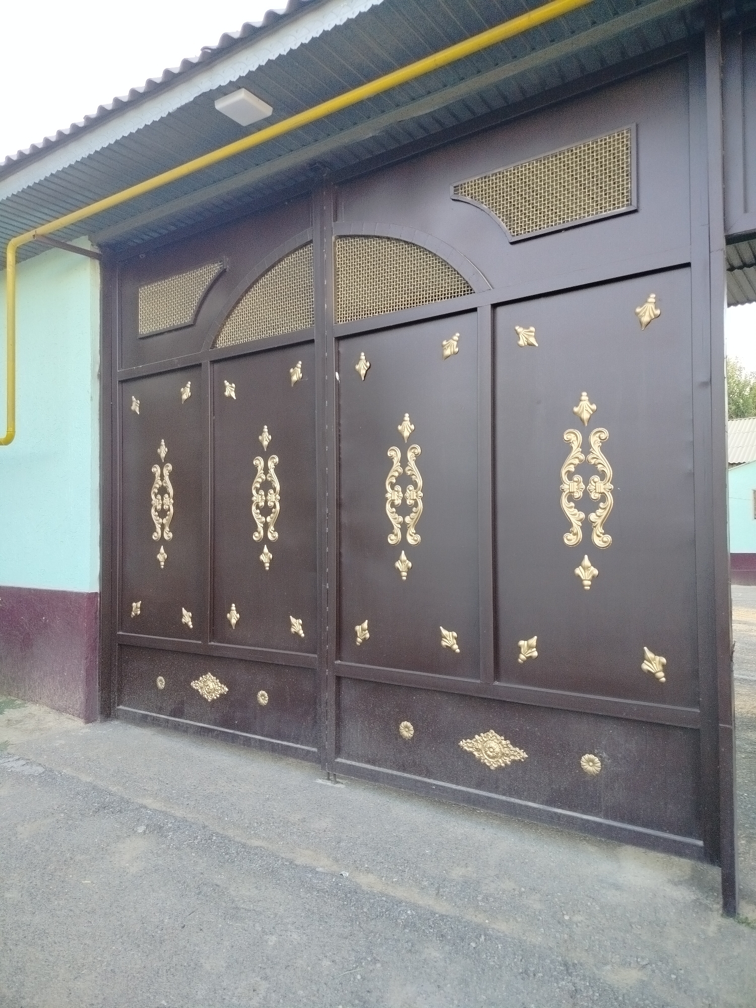 The image shows a dark brown metal gate with ornate gold detailing. The gate is divided into two large panels, each featuring intricate scrollwork and smaller decorative elements. The top section of the gate has arched openings covered with a gold mesh. Above the gate, a corrugated metal roof with a white trim is visible, supported by a light blue wall and a dark red base. A yellow pipe runs horizontally along the top of the gate and wall. The ground in front of the gate is paved with dark gravel. The scene is situated outdoors, with hints of greenery and a building in the background, suggesting a residential or commercial area. The architectural style and the presence of the yellow pipe may indicate infrastructure common in Central Asian cities like Көлкент, Kazakhstan. There are no people visible in the image.
