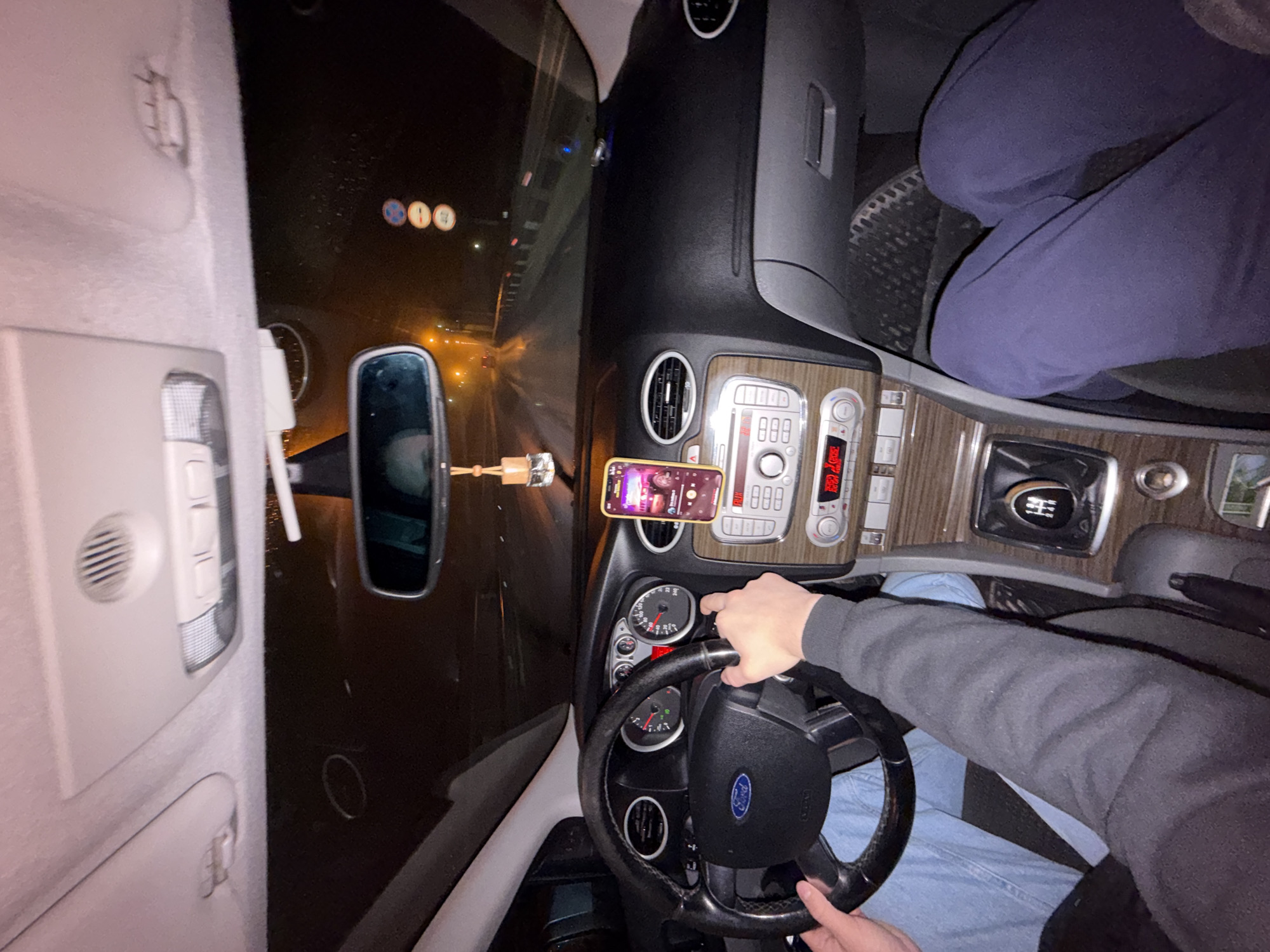 The image depicts the interior of a car, specifically a Ford Focus, at night. The dashboard, steering wheel, and front console are clearly visible. A mobile phone is mounted on the dashboard, displaying an app with a woman's picture visible. The driver's hands are on the steering wheel. Through the windshield, streetlights and road signs are visible, indicating the car is on a road. Based on the road signs, this is the city of Chaikovsky, Russia. There are two people in the front of the car, their legs are visible. The environment is dark, with the car's interior lights providing illumination. The scene appears to be a car in transit at night.