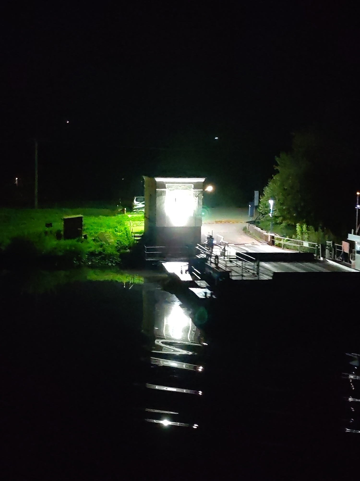 The image shows a nighttime scene of a cable ferry reflected in water, likely a river or canal. On the far bank, there's a small building emitting bright light, which is also reflected in the water. The surrounding area includes a grassy embankment and some trees on either side. The scene suggests it might be a rural or semi-rural location, and the cable ferry is the central element. The scene shows the Lesnoi cable ferry at night.
