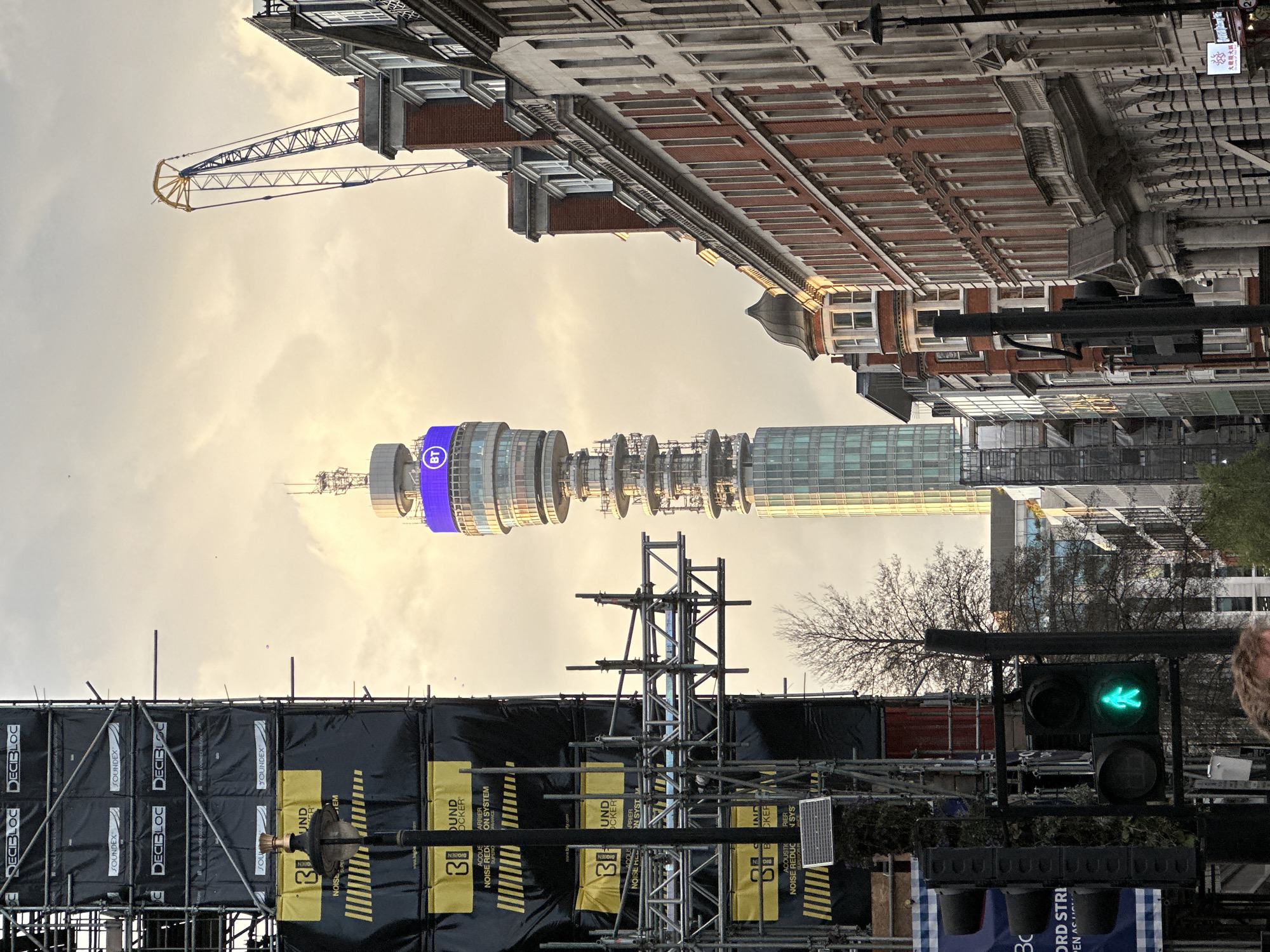 The image depicts a street scene in London, United Kingdom, with the BT Tower prominently displayed in the background. Scaffolding and construction materials dominate the foreground, including black barriers with yellow and white lettering. A traffic light shows a green walking symbol. The architecture includes traditional brick buildings and modern glass structures. A construction crane is visible above the buildings. The sky is overcast.