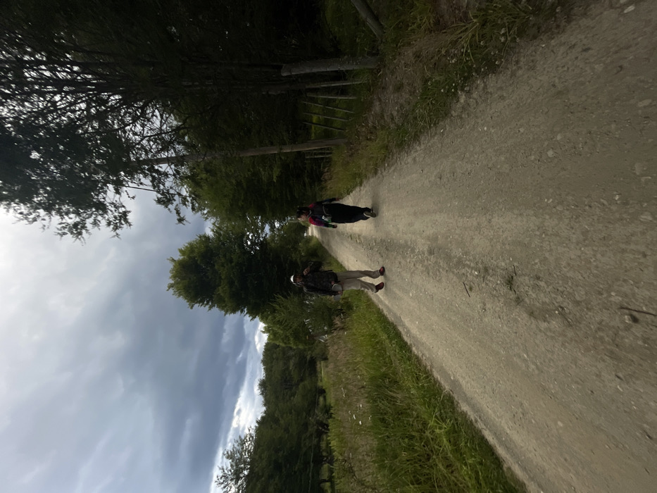 A dirt path stretches into the distance, flanked by trees and grassy areas on either side. Two people are walking along the path. The sky is partly cloudy. The photo appears to have been taken with a wide-angle lens and is rotated slightly. The lighting is somewhat dim, suggesting an overcast or late-day setting.