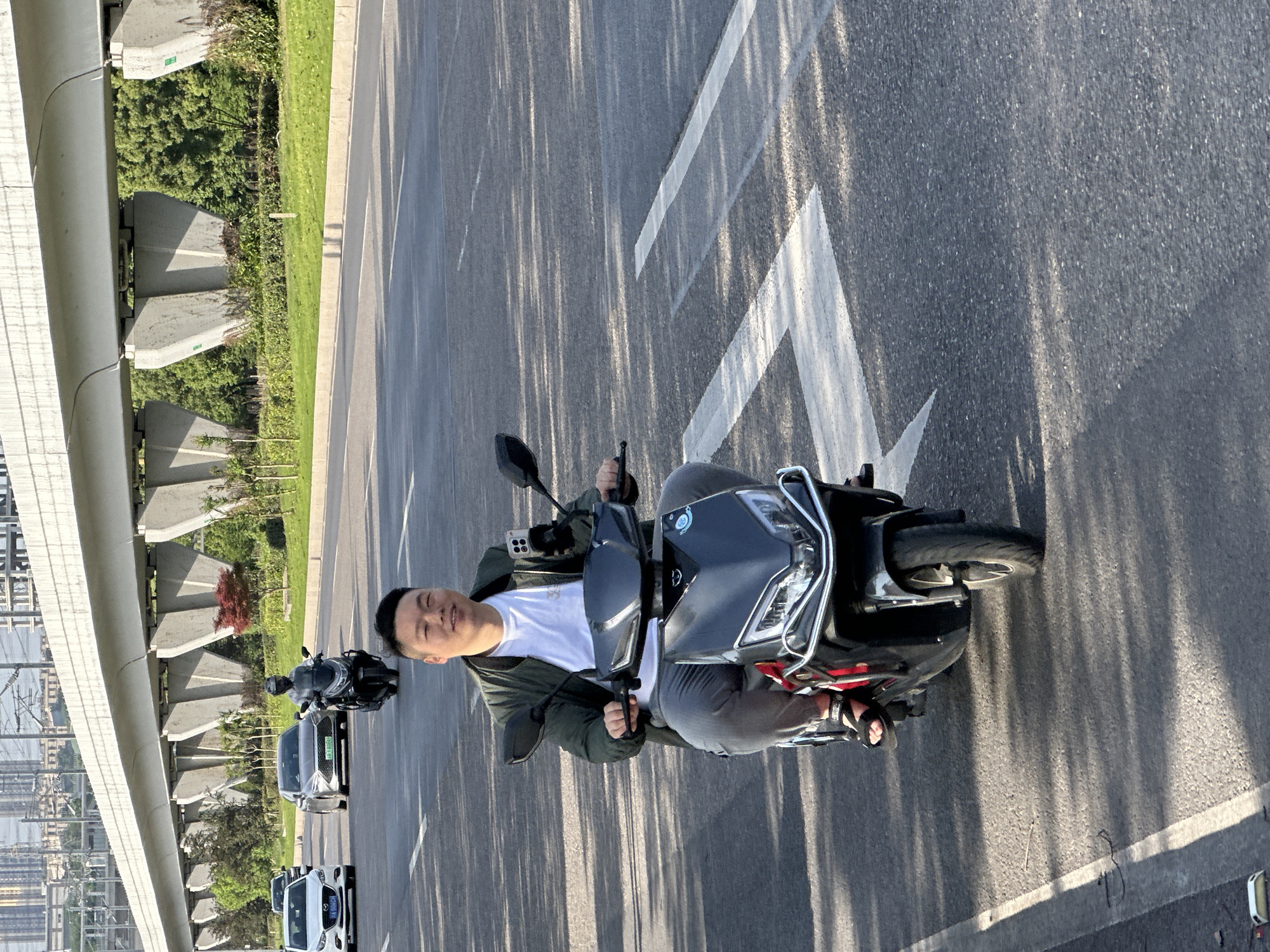 An outdoor, eye-level shot captures a man seated on a black electric scooter on an asphalt road in Jiangjin, China, bathed in bright sunlight. The man, wearing a white t-shirt, dark jacket, and dark pants with sandals, is smiling pleasantly towards the camera while holding the handlebars. An iPhone in a clear case is mounted on the scooter's handlebars.
The road features distinct white arrow markings and is streaked with long, narrow shadows, suggesting either morning or late afternoon sun. To the left, a prominent elevated concrete structure, likely a bridge or overpass, runs parallel to the road. Its supports are visible, nestled amidst green grass and dense foliage, including trees and bushes. In the background, beyond the greenery and bridge, more urban infrastructure such as power lines, pylons, and distant buildings can be seen.
Further down the road, behind the man, two cars are visible: a dark gray sedan with the license plate "渝AD 909C6" and a white SUV bearing the license plate "渝A 16K66". The overall scene depicts a typical urban or suburban setting on a clear, sunny day, with the man appearing relaxed and content.