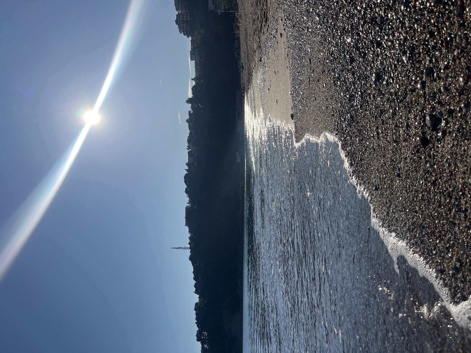 The image depicts a beach scene in Boreti, Montenegro. The sun is high in the sky, causing a bright glare and a long light streak. To the right, the shoreline is visible, composed of dark, pebble-like sand. White sea foam marks the edge where the water meets the sand. The water itself appears dark, reflecting the sunlight. On the left of the scene is a hillside covered in trees and sparse buildings. In the background, a tower-like structure can be distinguished amongst the trees. The sky is a clear blue.