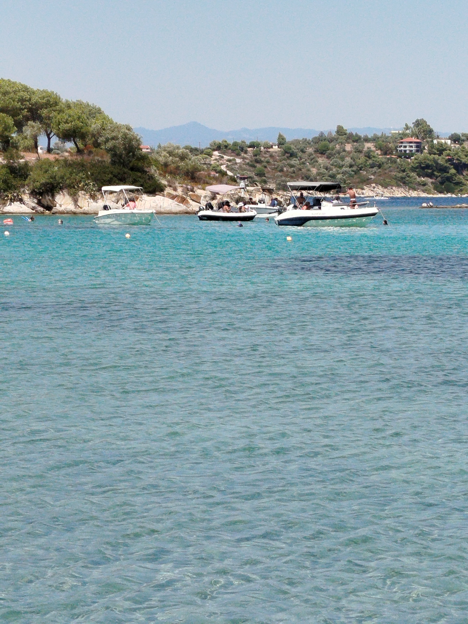 In the Sithonia Municipal Unit, Greece, a bright, sunny day is captured on the clear, turquoise waters of the Aegean Sea. The scene depicts a tranquil coastal environment with several boats anchored offshore, suggesting a popular recreational area.
In the mid-ground, a white motorboat with a blue stripe is prominently featured. It has a covered canopy and several people are seated on board. Another motorboat, also white, is visible to the left, with a similar canopy and passengers. In the distance, a third, smaller motorboat is partially visible.
The shoreline is characterized by rocky outcrops and lush green vegetation, including trees and shrubs. A few buildings, likely residences, are nestled into the hillside, overlooking the water. The water itself is calm, with gentle ripples disturbing the surface. A few buoys are scattered in the water, marking swimming or boating areas. The overall impression is one of a peaceful summer day at a seaside resort.