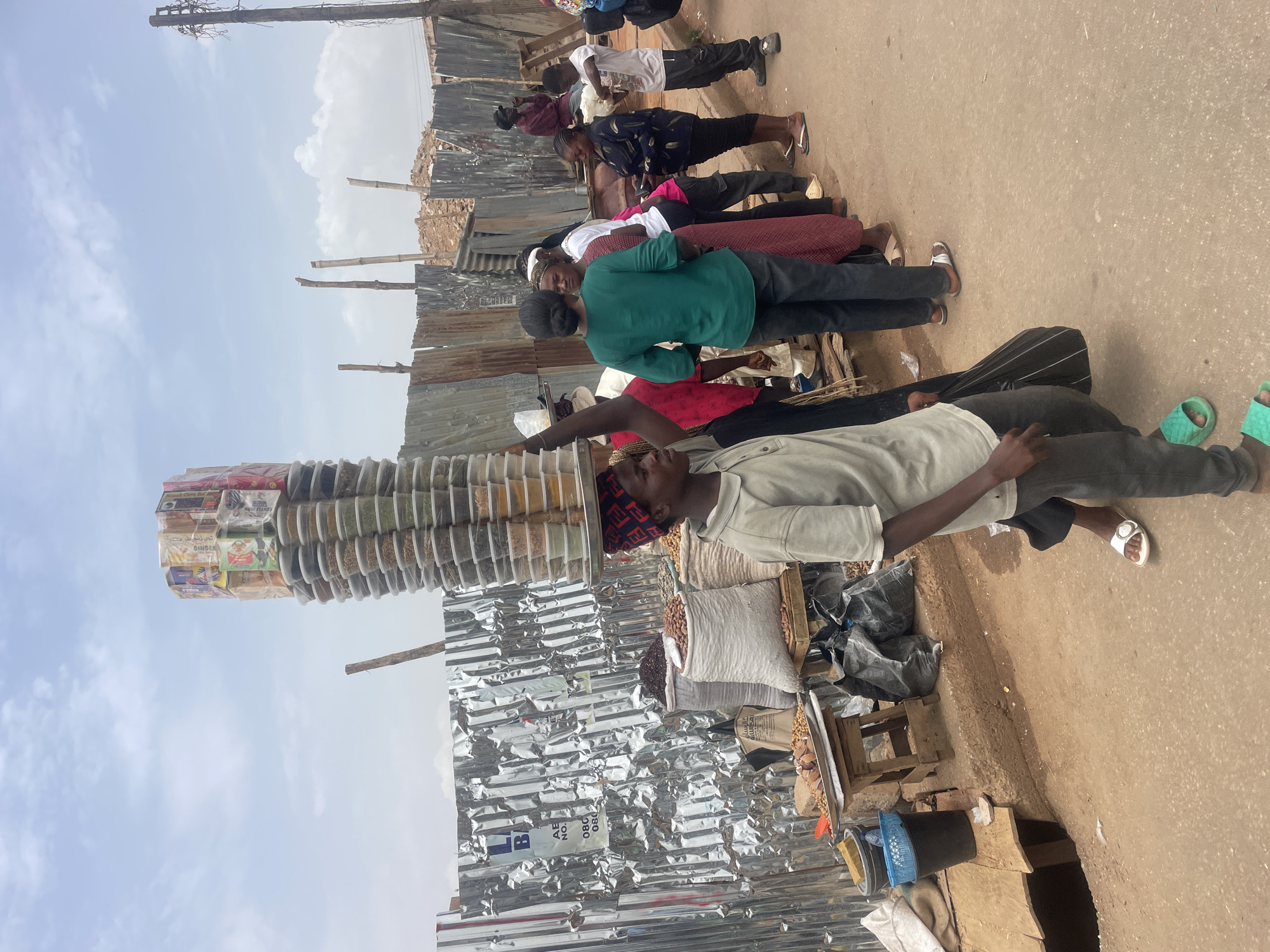 A street scene in Jos, Nigeria, shows a vendor balancing a tall stack of stacked plastic containers on his head. The containers, likely filled with spices, are layered atop each other. The vendor, wearing a light-colored shirt and dark trousers, stands near his stall. The stall displays bags of items. A line of people standing nearby appear to be customers. The background features corrugated metal structures. The street is unpaved and dusty. The sky is overcast.