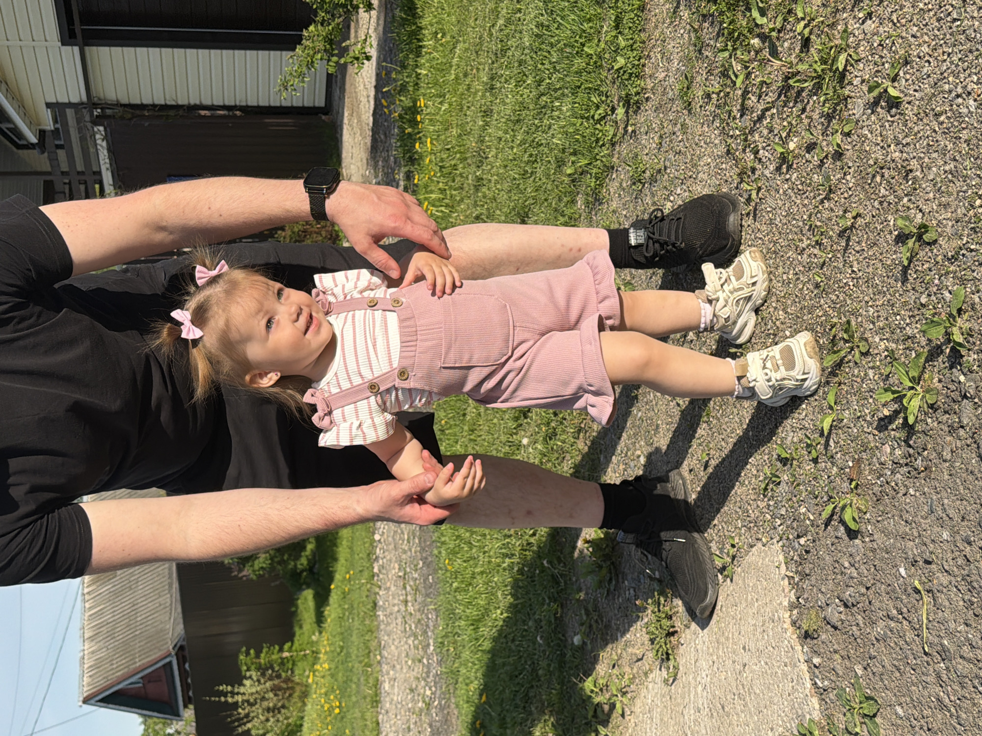 A young girl in a pink romper with pink bows in her hair is held supine by an adult man in a black shirt and dark sneakers. The man is grasping the girl's hands and also supporting her torso with his other hand. The location is outdoors, likely a yard or park in Reshetnikovo, Russia, evidenced by the presence of greenery, gravel, and glimpses of residential buildings.