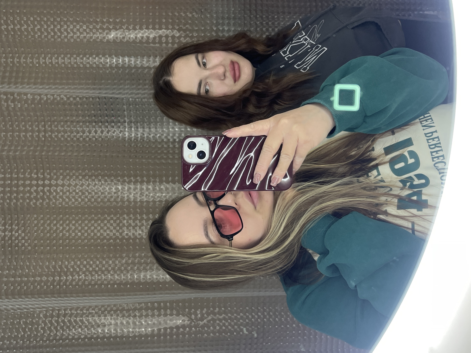 Two women are visible in the image, seemingly taking a selfie in front of a mirror. One woman has brown hair and is wearing a dark top with white writing. The other woman, holding the phone, has blonde highlighted hair and is wearing red sunglasses and a green sweatshirt with a light green square design on the sleeve. The phone being used for the selfie has a purple case with white stripes. The background appears to be a corrugated metal surface, with a reflection suggesting the location might be in Krasnodar, Russia due to the visible Cyrillic text in the reflection. The women are looking at the camera, and the interaction is a casual selfie-taking activity.
