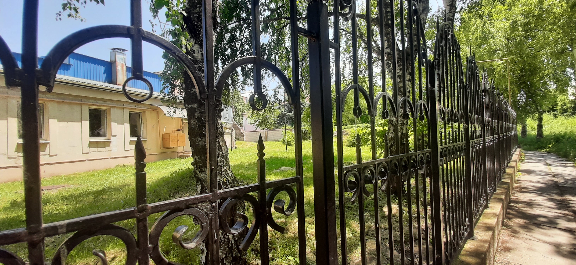 An exterior shot captures a sunlit scene featuring a black wrought-iron fence in the foreground, partially obscuring a light yellow building with three windows and a blue-roofed structure behind it. The fence, characterized by decorative scrollwork and vertical bars, runs along a paved pathway bordered by a low brick wall. Beyond the fence and building, a grassy area is visible, with several trees, including a prominent birch tree with striking white bark, interspersed with lush green foliage. The sky is clear and blue. The image appears to be taken in Yessentukskaya, Russia, given the architectural style of the building and the surrounding vegetation typical of the region. There are no people or animals present in the scene.