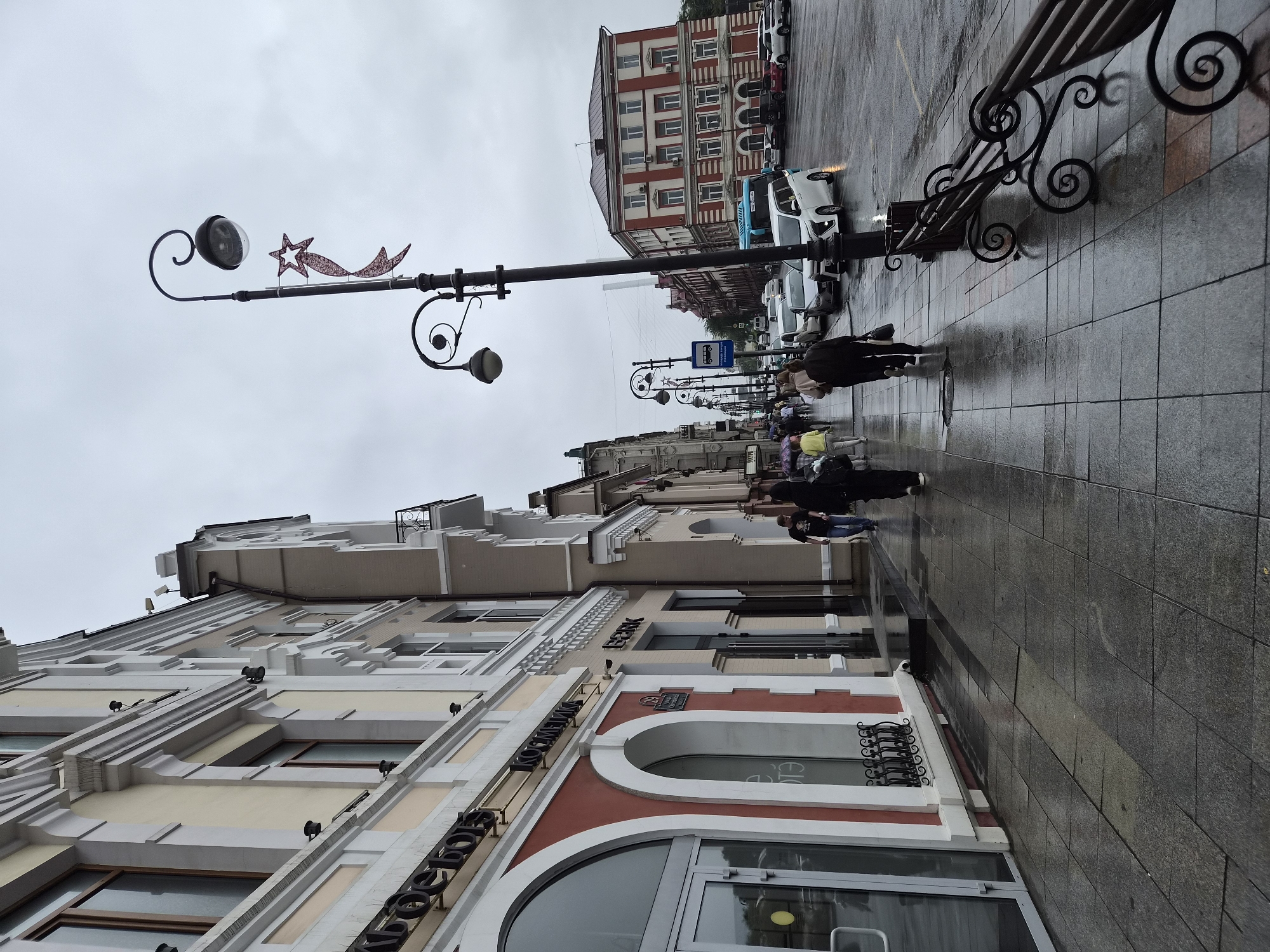 The image depicts a street scene in Vladivostok, Russia, on a cloudy day. The wet pavement indicates recent rain. On the left, a row of buildings with cream and red facades lines the sidewalk. Several storefronts are visible, one with large arched windows and signs including "Косметик". Pedestrians walk along the sidewalk.
Further down the street, a distinctive red and white building stands on the right. Traffic, including cars and a blue bus, moves along the wet road. Ornate streetlights with decorative elements, including a star and ribbon, are spaced along the sidewalk. A wrought-iron bench sits on the right side of the sidewalk. The overall atmosphere is one of a typical urban street on a damp day, with architectural details characteristic of Russian cities.