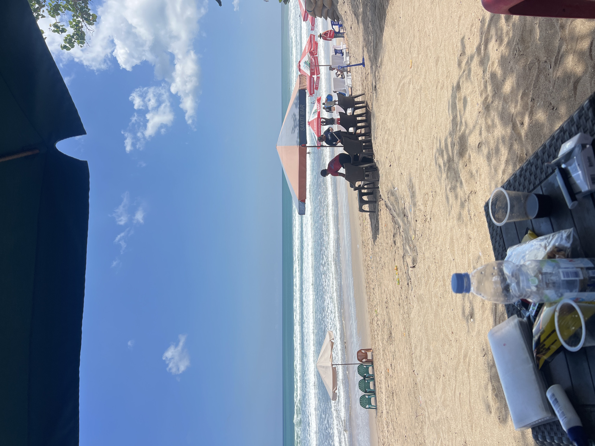 This is a photograph of Kuta Beach in Kuta, Indonesia, on a sunny day. The foreground shows a table with various items on it, including a bottle of water, a pack of cigarettes, and a plastic cup with a dark liquid. The table is made of woven material and is positioned on the sand. Beyond the table, the sandy beach stretches out towards the ocean.
Several beach umbrellas are set up along the shore, some with chairs and tables beneath them. In the middle ground, a group of people are gathered under a large orange and gray umbrella. Some appear to be setting up chairs, while others are conversing. One person in a red shirt is arranging chairs. Further down the beach, more umbrellas and beachgoers are visible.
The ocean is a prominent feature of the image, with gentle waves rolling onto the shore. The water is a turquoise blue, and the sky above is clear with scattered white clouds. A few small, white clouds are present in the distance, and a larger tree branch with green leaves extends from the right side of the frame, casting a shadow on the sand. The overall scene depicts a relaxed atmosphere at a popular tourist destination.