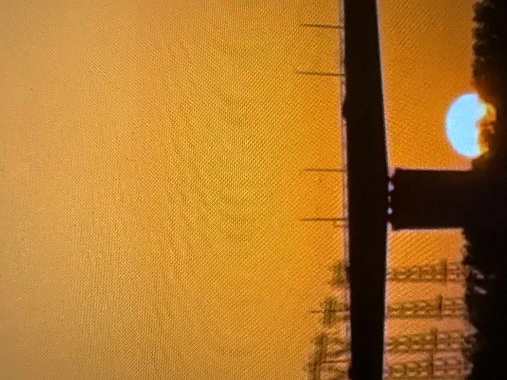 The image shows a sunset in an urban environment. A utility pole is visible in silhouette, with attached wires and equipment. The sun is a bright white-blue orb, partially obscured by the pole and what appears to be a building or other structure. The sky is a uniform orange color, suggesting either a sunset or heavy air pollution. The details of the building are obscured by the darkness. The city name and location are not determined due to the limited visual information.