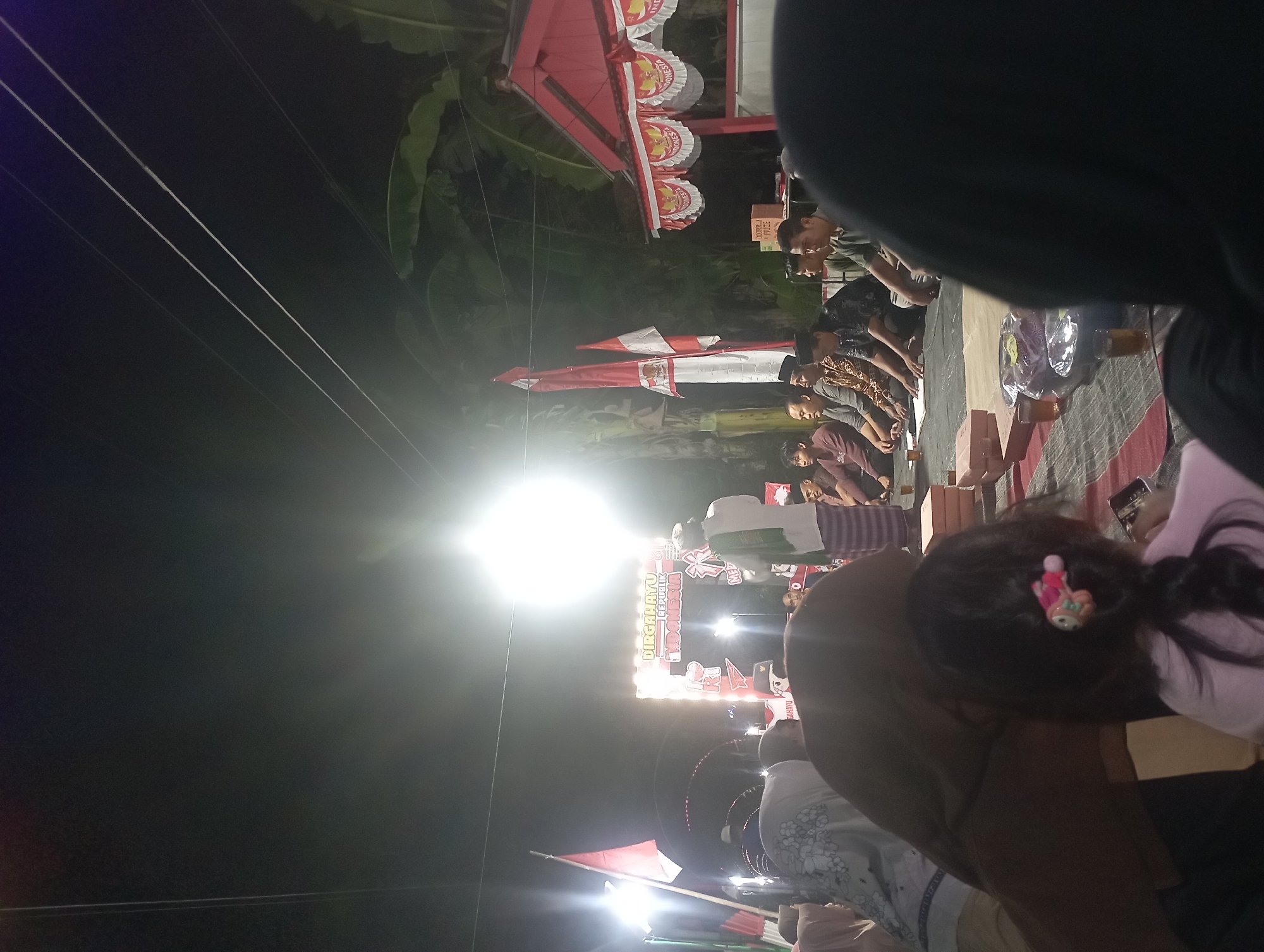 This is a night-time outdoor scene in Maos Kidul, Indonesia, with a gathering of people sitting on mats. The event appears to be a celebration or cultural gathering, indicated by Indonesian flags and banners reading "Dirgahayu Republik Indonesia." A brightly lit archway with the same inscription serves as a focal point. Several men are seated in the foreground, some wearing traditional attire. A man in a white shirt and green scarf stands near the archway, possibly addressing the crowd. In the background, there are trees, a small red building with white trim and Indonesian flags draped around it, and power lines overhead. The atmosphere is illuminated by strong artificial lighting, creating a festive and community-oriented environment.