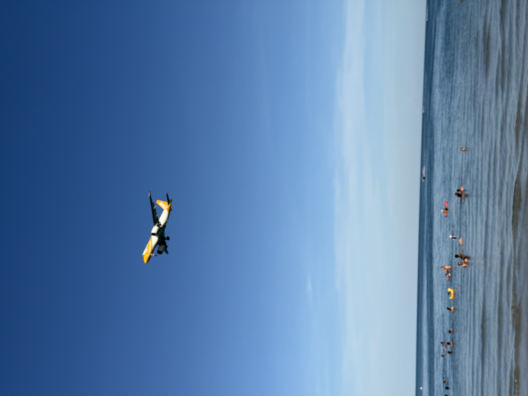 An aerial view shows a yellow and white airplane flying against a clear blue sky. Below the airplane, a body of water, likely the ocean, is visible. Several people are scattered throughout the water, some swimming and others relaxing on inflatable objects. A small yellow duck-shaped inflatable is present. The water appears calm with gentle ripples. In the distance, the horizon line separates the water from the sky. The scene depicts a typical beach day with recreational activities in progress. The specific location is Baan Chaweng, Thailand, known for its beaches and tourist activities.
