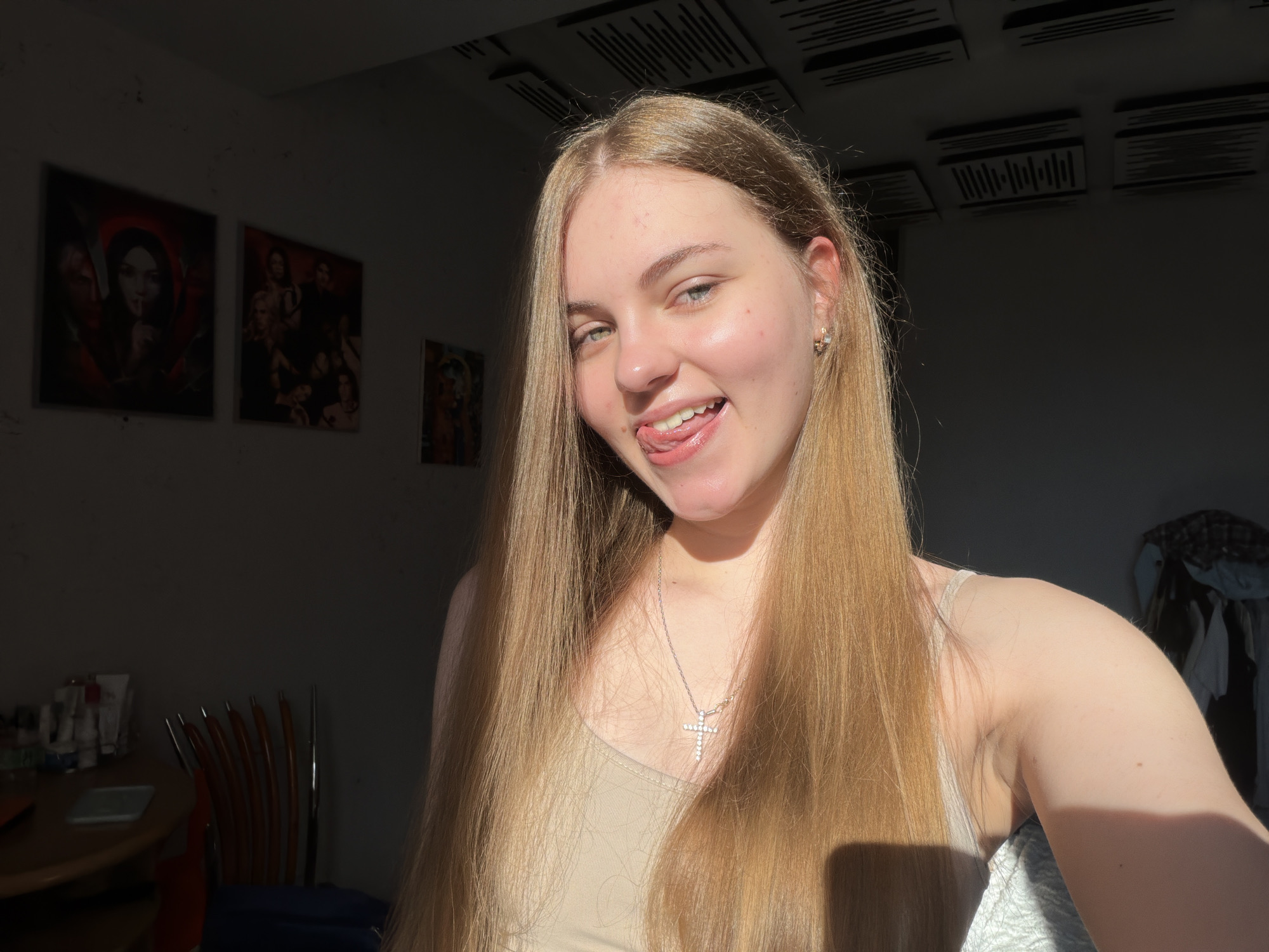 A young woman with long, blonde hair is shown from the chest up, facing the camera. She is sticking her tongue out and winking. She is wearing a light-colored tank top and a silver necklace with a cross pendant. The background is a dimly lit room with several framed pictures on the wall. The room appears to be a bedroom or living area. The woman's face is illuminated by sunlight coming from the left side of the frame.