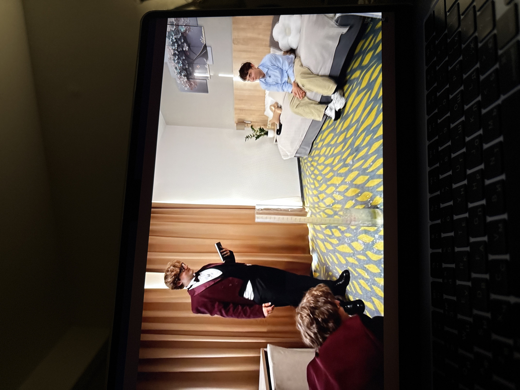 This image shows a laptop screen displaying a video. On the screen, in what appears to be a hotel room, a young man dressed in a maroon tuxedo jacket, white shirt, and black pants is standing and holding a black book. Another person, whose back is to the camera and wearing a maroon garment, is leaning over in the foreground. In the background, a second young man is seated on a bed, wearing a light blue collared shirt and khaki pants. The room has a patterned yellow and blue carpet, a bed with light-colored bedding, and a wall with a framed artwork depicting a tree with flowers. The image does not provide any specific details to indicate a location in the city of Perm, Russia.