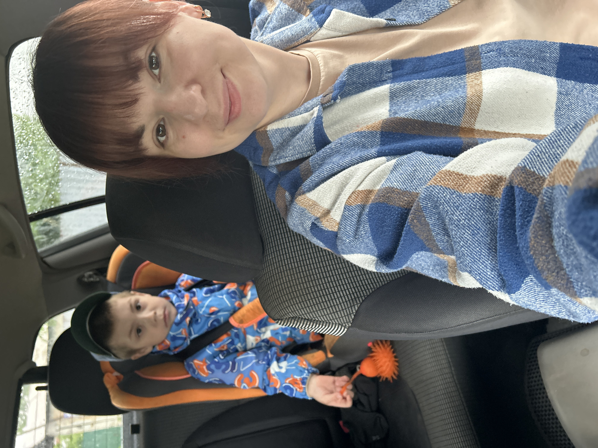 The image shows a woman and child in a car. The woman is in the foreground, facing the camera. She has short brown hair and is wearing a blue and white plaid shirt. Behind her is a car seat with a child wearing a blue jacket with orange designs and a green cap. The child is holding an orange spiky ball. The scene is inside a car, and it appears to be raining outside, based on the raindrops visible on the window. The location is Фоки, Russia.