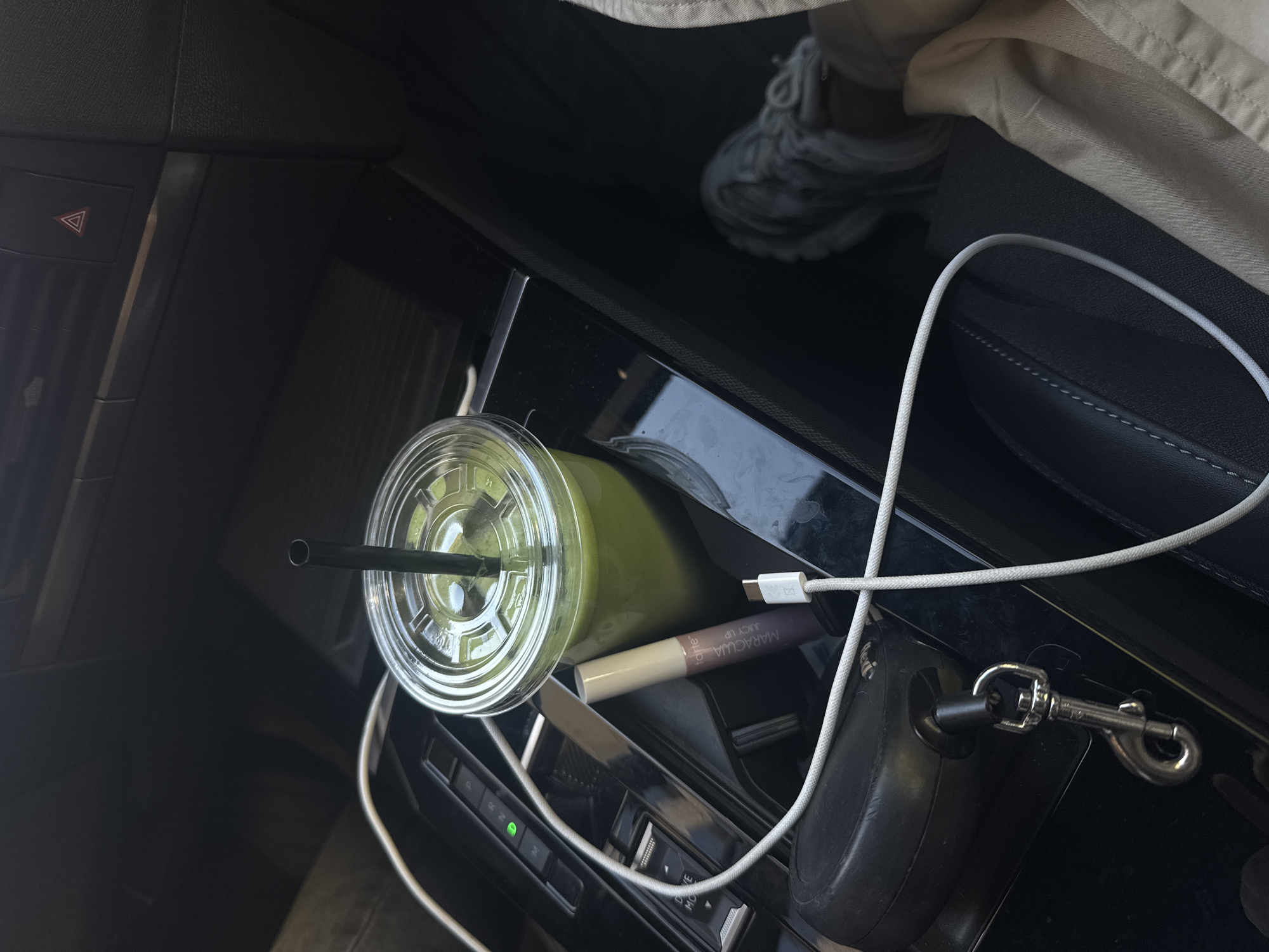 This image presents a close-up, high-angle view inside a car, with the central console and adjacent seating area as the primary focus, set in Warsaw, Poland.
In the foreground and center, a clear plastic cup containing a vibrant green beverage, likely a smoothie or juice, is positioned in a cup holder. A black straw protrudes from the clear lid. A white charging cable snakes across the console, connecting to a port near the drink. Adjacent to the cup, a light beige cosmetic tube is visible, clearly showing the brand name "tarte" and the product name "MARACUJA JUICY UP". Further to the right, a silver carabiner-style clip, typically used for a dog leash, is attached to a black car accessory, possibly a retractable leash housing or a storage hook.
To the left, car controls are visible, including a prominent hazard warning light button with its characteristic red triangle symbol. Further down the console, gear selector or drive mode controls are present, with the text "DRIVE MODE" faintly discernible and a small green indicator light illuminated. A person's leg, dressed in light-colored trousers, and a grey athletic shoe are partially visible in the upper right background, suggesting a driver or passenger occupies the car. The car's interior features dark surfaces, including stitched upholstery on the seat. The lighting indicates it is daytime. The scene depicts everyday items commonly found within a vehicle, suggesting a moment of travel or a stop.