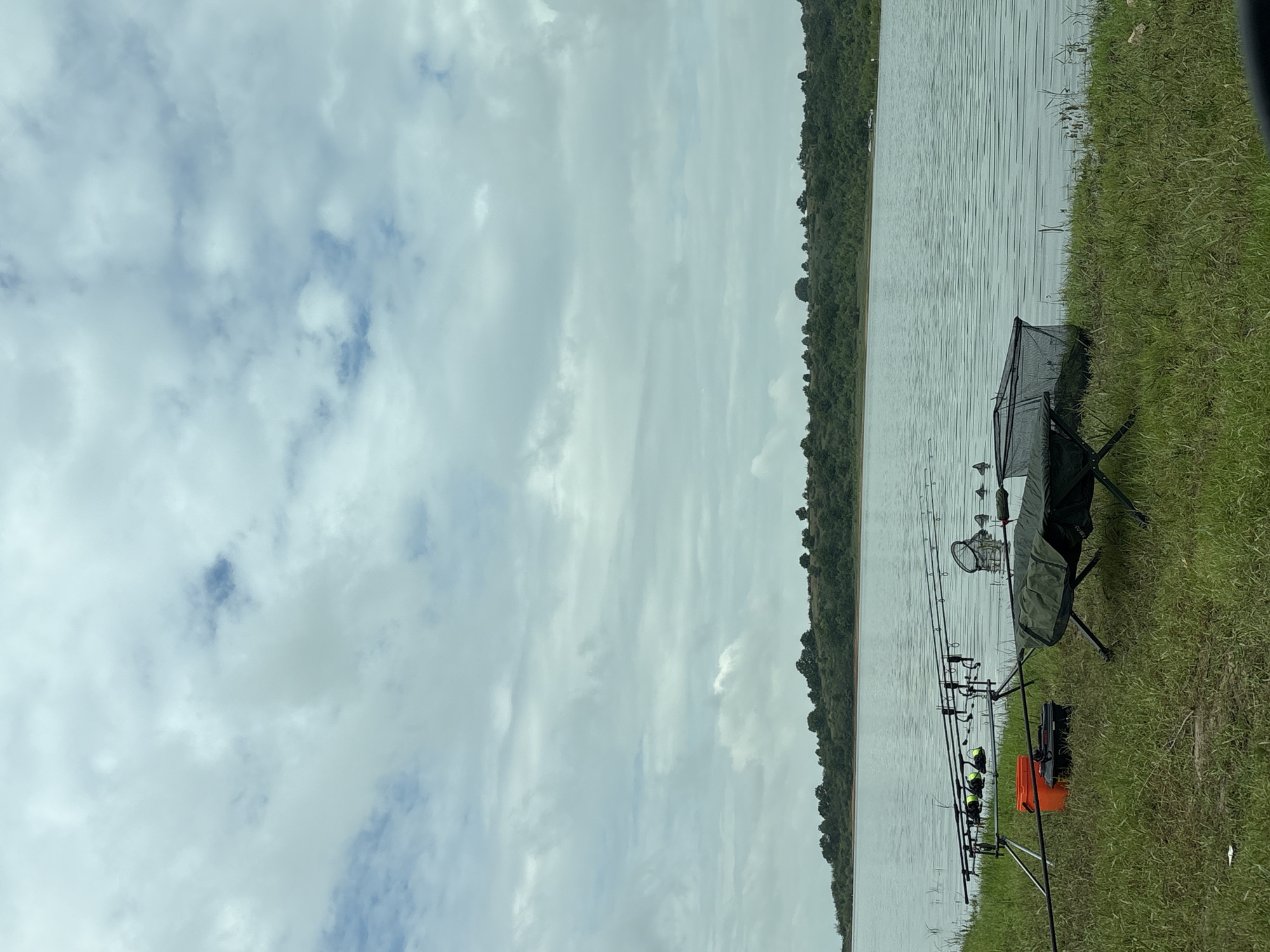 The image shows a fishing scene at a body of water, possibly a lake or river, near Brezovo, Bulgaria. Several fishing rods are set up on a rod pod with electronic bite alarms attached. A green chair and a keepnet are positioned near the fishing rods. An orange bucket is visible, likely containing fishing tackle or bait. The shoreline is grassy. Across the water, a line of trees is visible. The sky is overcast with grey and white clouds.