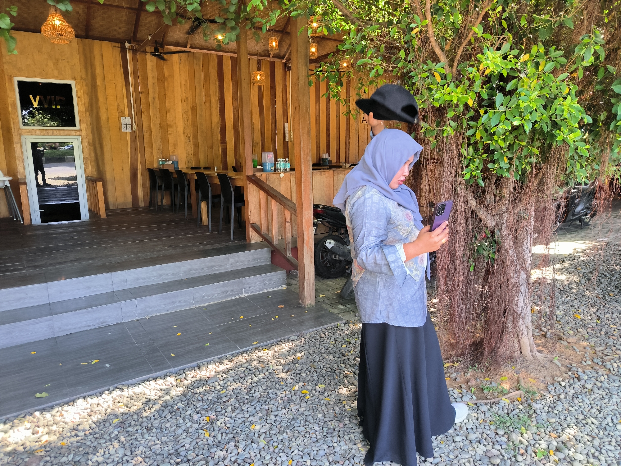 A woman wearing a light blue hijab and a long black skirt is standing outside a wooden building with a gravel ground in Santan, Indonesia. She is holding and looking at a purple smartphone in her hands. A black hat is being held above her head. The building has a sign that says "VVIP" and there are tables and chairs inside. A tree is next to the woman. A motorcycle is parked near the building.