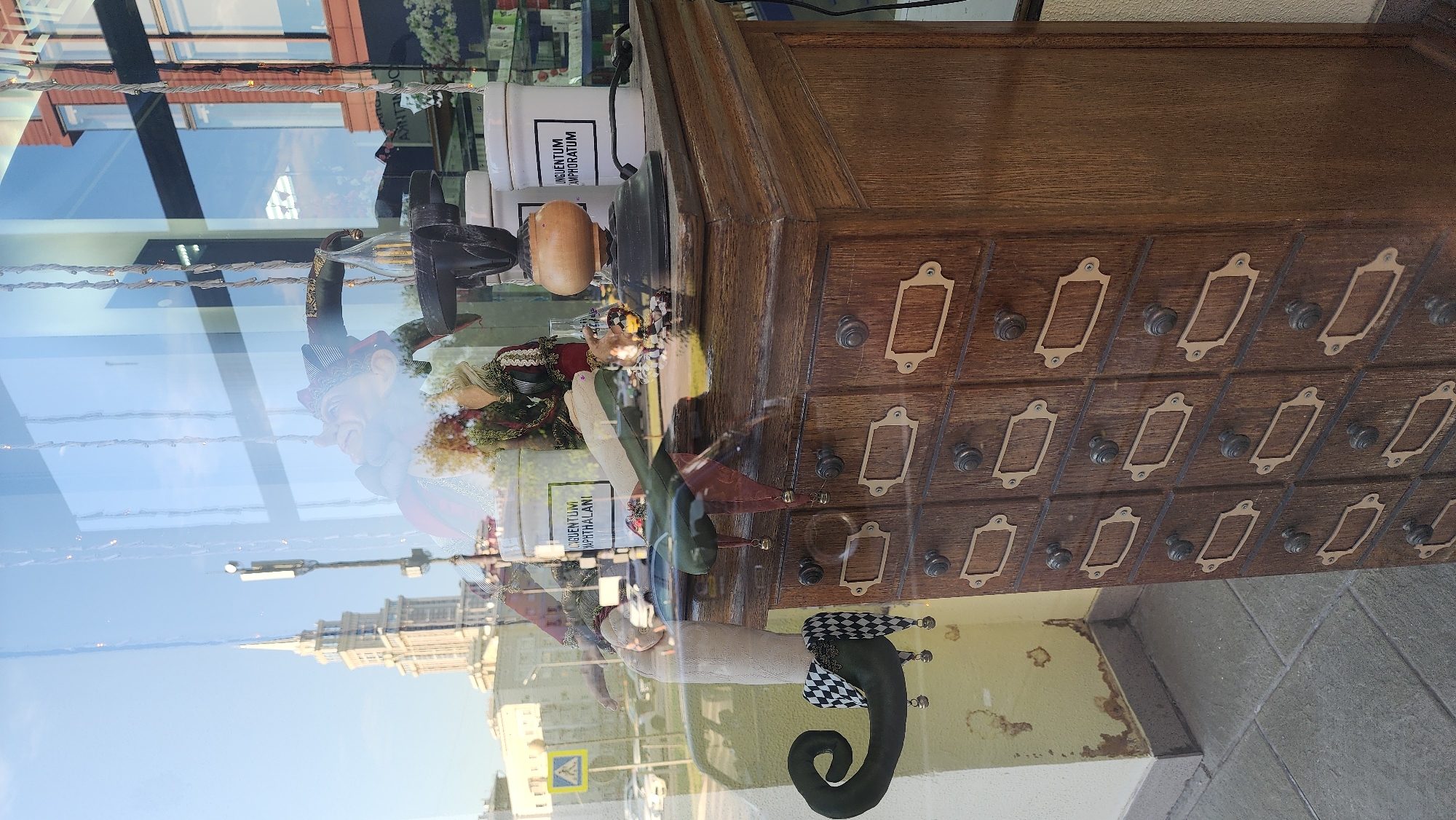 A reflection in a shop window shows a multi-story building with a spire, resembling a Moscow Stalinist skyscraper, against a bright blue sky with scattered clouds. A yellow pedestrian crossing sign is visible on the left. Inside the shop, a wooden cabinet with many small drawers is displayed. A whimsical figure with a jester's hat and a long, pointed green shoe with bells is placed to the left of the cabinet, partially obscuring a white jar with text. A vintage-style lamp with a wooden base and a dark metal shade sits on top of the cabinet, next to another white jar with text. The reflection on the glass distorts the view of the items within the shop, creating a layered effect with the external environment.