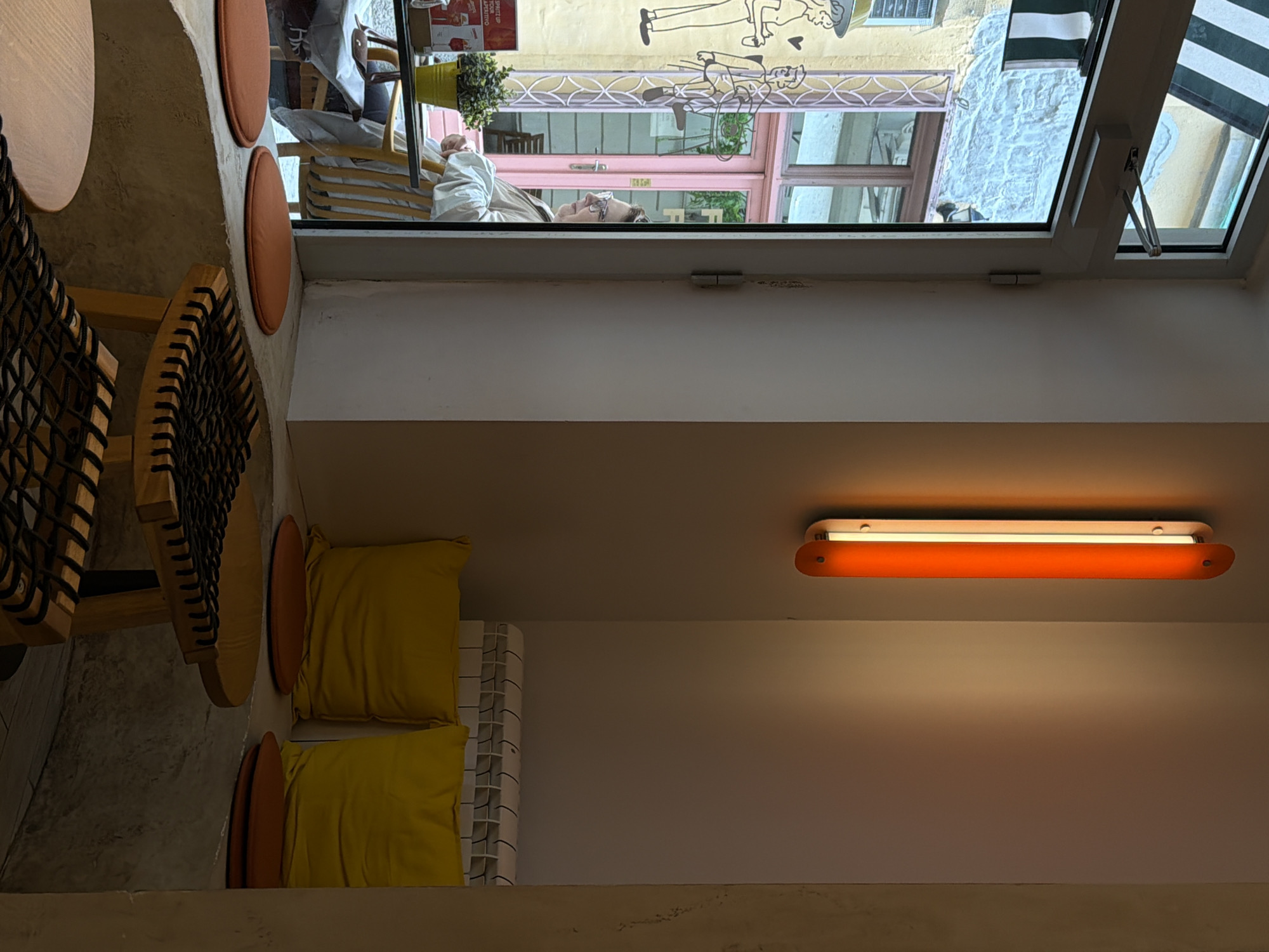The image captures an interior scene, seemingly in a café or small shop, possibly in Palilula Urban Municipality, Serbia, based on the architectural style reflected in the window view. A wooden chair with a woven seat is positioned near a wall, accompanied by yellow pillows. A warm-toned, elongated light fixture is mounted on the wall. Through the window, one can glimpse a street scene with pastel-colored buildings, people, and decorative elements like a flower pot. The viewpoint is from within the space, looking out onto the street. A person's face can be seen reflected in the window.