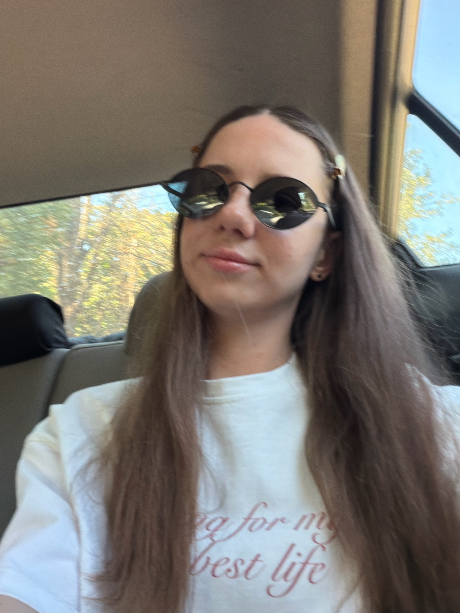 A young woman with long, brown hair, wearing sunglasses and a white t-shirt, is seen from the chest up, inside a vehicle. The t-shirt has pink text on it, which appears to say "ing for my best life". The background shows trees and a blue sky through the car window, suggesting an outdoor environment. The image appears to be taken with a handheld camera, possibly a selfie. There is no indication of the location being Kyiv, Ukraine.