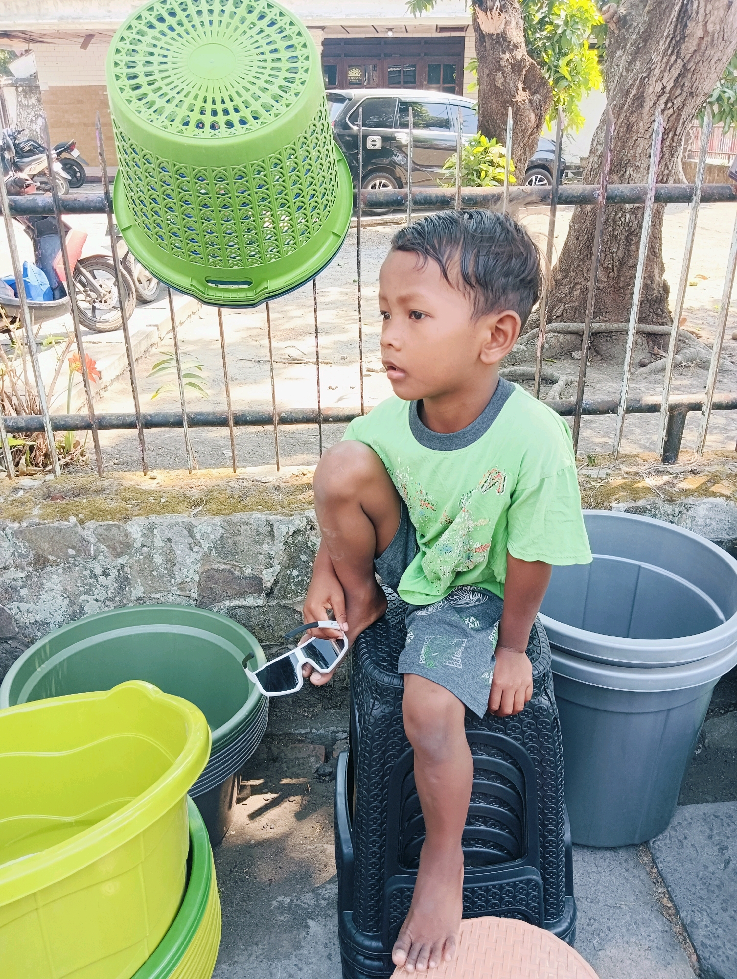 In Ploso, Indonesia, a young boy, appearing to be around 5-7 years old, sits on a black plastic stool in what seems to be a residential outdoor area. He is wearing a light green t-shirt with a graphic print and gray shorts with a patterned design. He is holding a pair of white-framed sunglasses in his right hand, and his legs are bent, with his left knee raised.
Surrounding him are several large plastic containers. To his left, two green and lime green tubs are stacked. To his right, a large gray tub is visible. Behind him, a metal fence runs horizontally, with what appears to be trees and parked vehicles visible in the background. A large green laundry basket is hanging from the fence above and to the left of the boy. The ground appears to be a mix of dirt and pavement. The lighting suggests it is daytime, likely sunny.