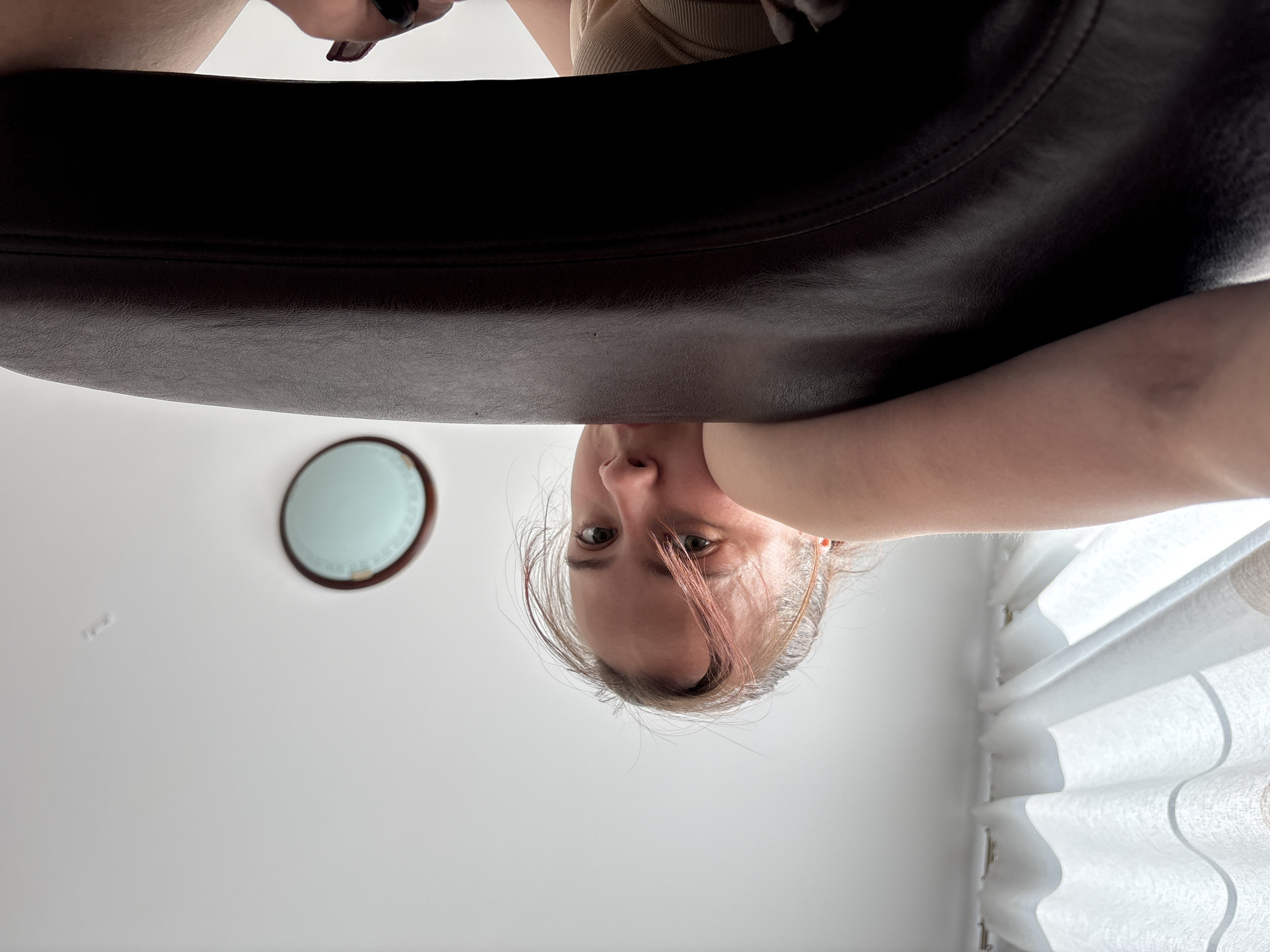 The image is an upside-down shot taken from a low angle. A person with light skin, blonde hair with pink highlights, and visible eyes and nose is in the lower center. Above them is a dark brown leather surface, possibly the armrest of a chair. In the upper left is a partially visible light fixture on a white ceiling. To the right of the person are white curtains. There is no information to suggest the image was taken in Perm, Russia.