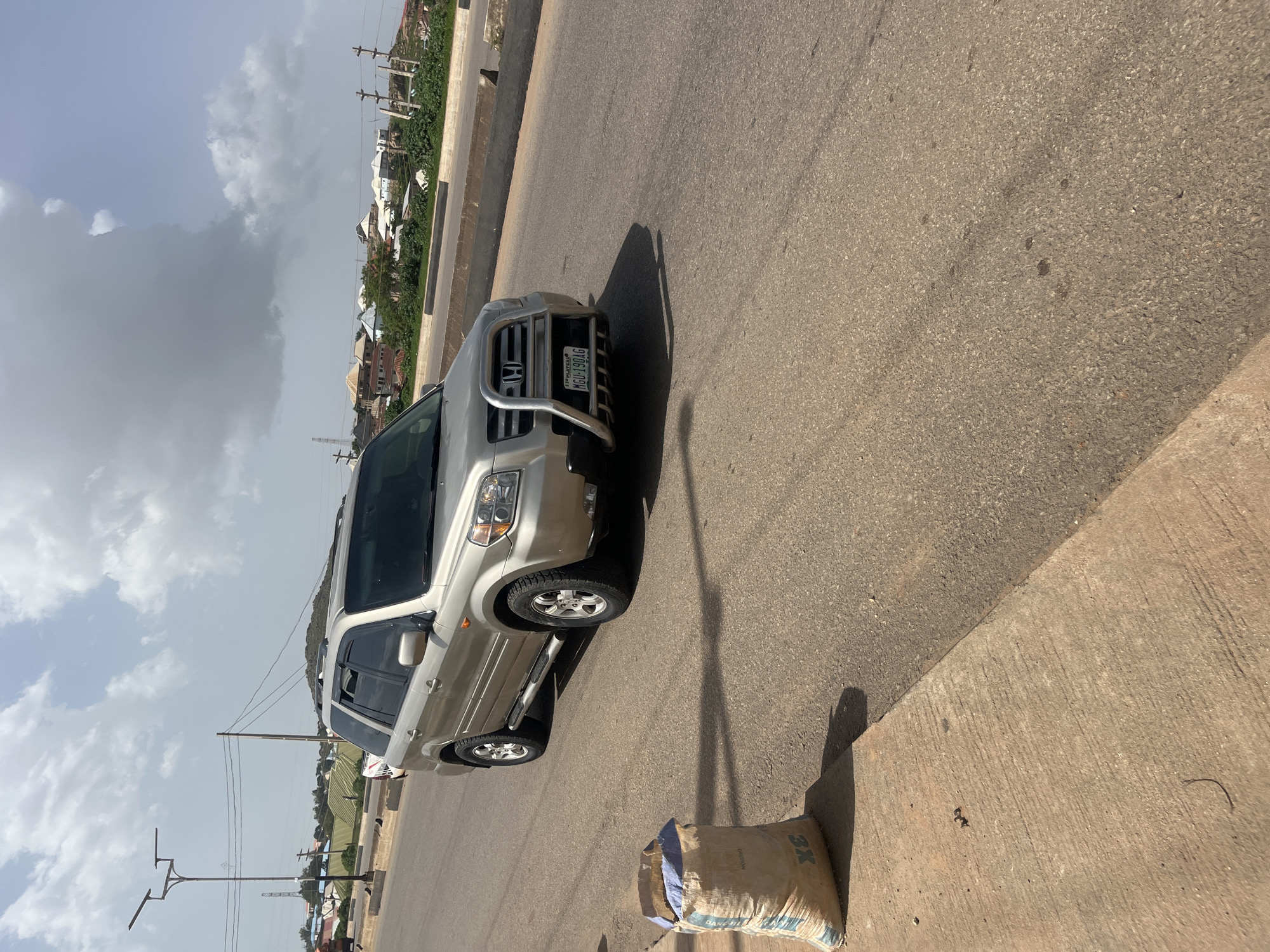 The image depicts a light gold Honda Pilot SUV parked on a paved road in Gura Top, Nigeria. The vehicle is facing slightly towards the camera and is positioned on the right side of the road. It has a silver bull bar with a Honda emblem on the front. The license plate reads "MGU-190AG". To the left of the vehicle, on the concrete sidewalk, is a large, light-colored burlap bag with blue and white markings and the text "CAUTION" visible on its side, along with "3X". In the background, there are buildings with pitched roofs, some greenery, utility poles with power lines, and a street lamp with multiple fixtures. The sky is partly cloudy with large, white and grey clouds. The scene appears to be a street in a semi-urban or developing area.