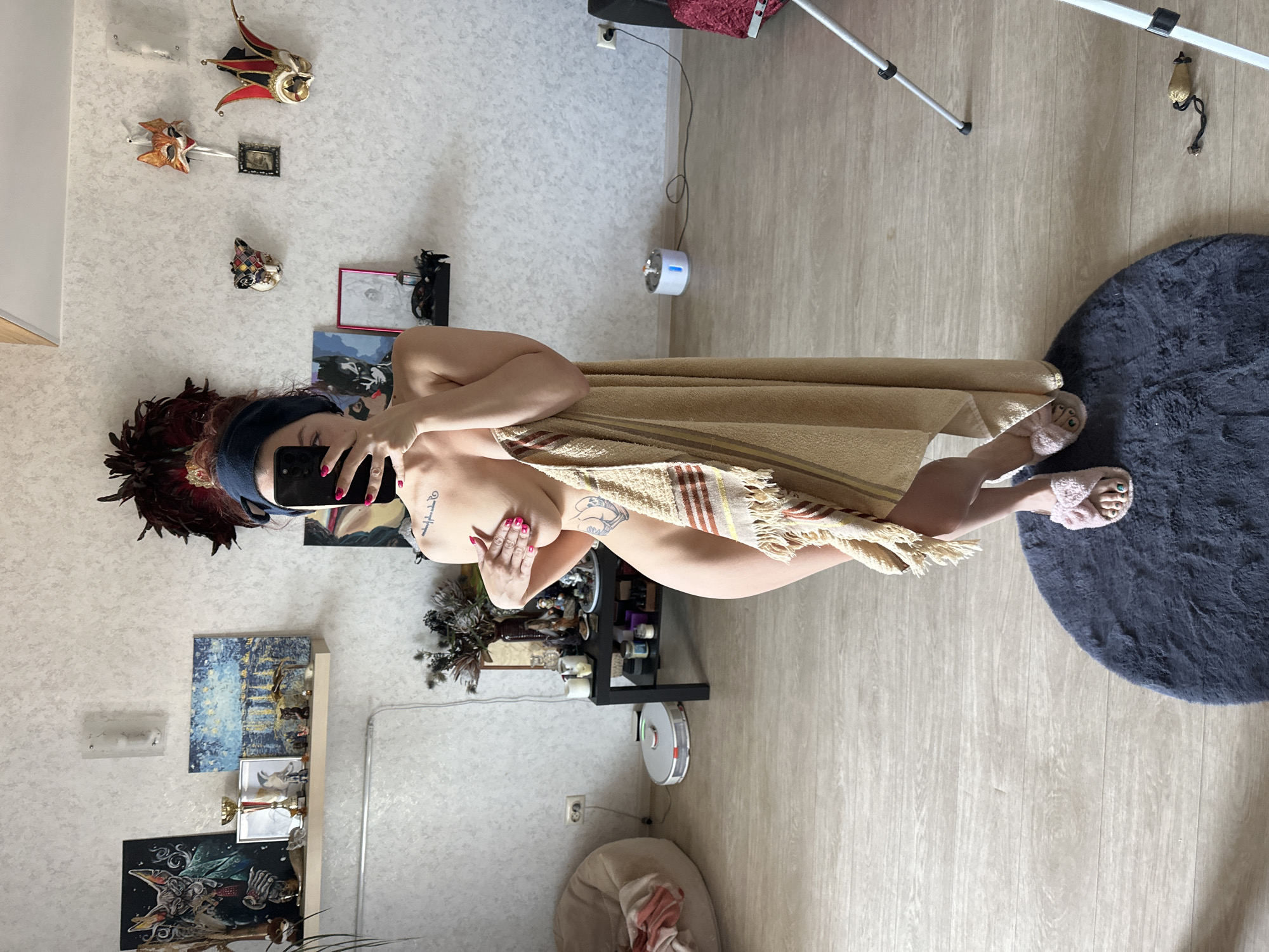 A female individual is depicted standing in what appears to be a residential interior, located in the city of Vladimir, Russia, taking a mirror selfie with a black smartphone. Her head is wrapped in a dark blue fabric. She is partially clad, with a tan-colored towel covering her torso and lower body, held by her left hand. Her right hand, with pink-painted fingernails, holds the smartphone towards her face, partially obscuring it. A tattoo of text is visible on her left clavicle/shoulder area. Another tattoo, resembling a stylized heart or figure, is visible on her left hip/thigh. She is wearing pink, fuzzy slippers with bows, and her toenails are painted green. She stands on a dark grey, semi-circular shag rug, positioned on a light-colored laminate wood floor.
The background consists of a light-colored, textured wallpaper. To the left, a white floating shelf displays a framed artwork featuring a stylized cat-like figure in a suit, a golden trophy cup, a smaller framed picture, several small figurines, and a painting with blue and yellow tones depicting a city scene at night. A wall sconce is mounted above this shelf. A trailing plant is visible behind the shelf. A beige bean bag chair with a folded blanket rests on the floor to the far left. Next to it, a black rectangular table holds a dark floral arrangement in a vase, candles, and various small objects. A white and grey circular robot vacuum cleaner is positioned on the floor in front of the table.
To the right, a series of decorative masks are mounted on the wall, including a multicolored harlequin mask, a fox mask, a red, black, and gold jester hat-like mask, and a partially visible white mask. A white pet water fountain with a blue light is on the floor, connected by an electrical cord to a wall outlet. A black speaker or electronic device is situated on the far right, next to a silver-colored tripod. A small, light-colored object with a black string lies on the floor near the base of the tripod.