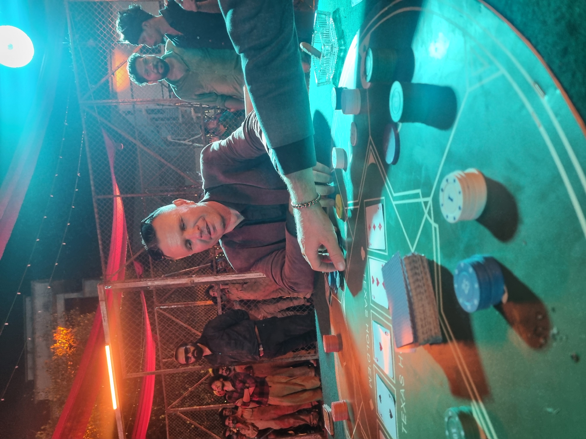 The media file captures an evening scene in Kalamassery, India, depicting a lively card game, likely poker. The central subject is a man with dark hair and a light complexion, wearing a dark jacket and a silver beaded bracelet, who is leaning over a round, green Texas Hold'em poker table. He is looking towards the viewer with a neutral expression, his hands positioned near poker chips on the table.
The table, clearly marked with "TEXAS HOLD'EM" text in white, is laden with stacks of poker chips in various colors (white, blue, red) and several playing cards. A clear glass ashtray is also visible on the table. In the background, several other men are standing, observing the game, some with beards.
The setting appears to be an outdoor or semi-outdoor event, with a chain-link fence and metal scaffolding visible in the background. The scene is illuminated by striking artificial lighting: a prominent, circular blue-green light source on the left casts a strong glow, and several linear orange or red lights create warm accents. Red fabric drapes are also seen hanging in the background. The time of day is clearly night, as indicated by the reliance on artificial lighting. The overall atmosphere suggests a social gathering or a game night.