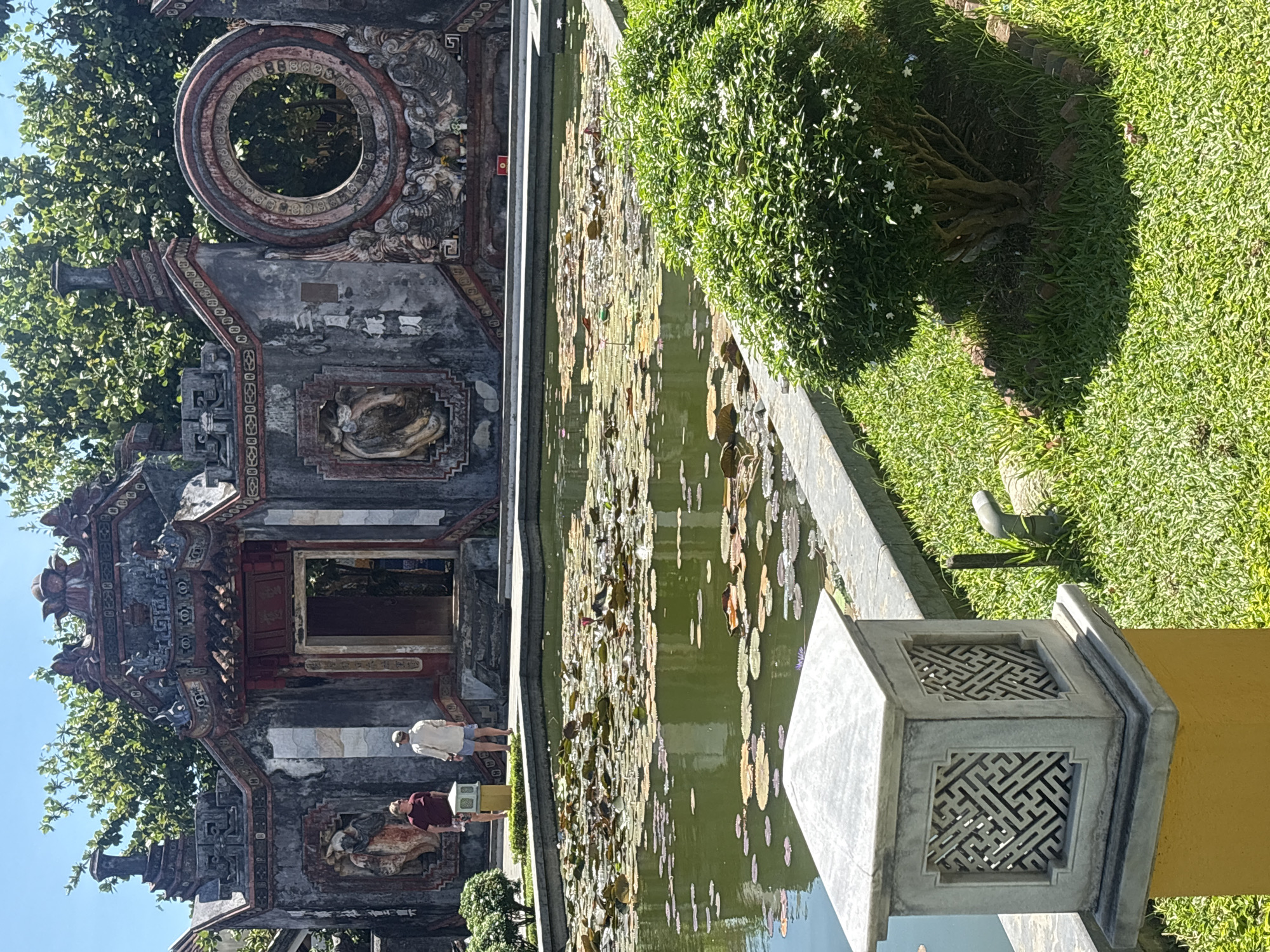 The image shows a serene courtyard with a pond filled with lily pads. In the background, an ornate, ancient structure with intricate carvings and a circular window stands against a clear blue sky. Two individuals, a man in a maroon t-shirt and shorts and another in a cream-colored shirt and blue shorts, stand on a stone pathway. A decorative lamppost with a geometric design is in the foreground, next to a lush green bush. The scene is characteristic of the historic gardens and temples found in Hội An Ward, Vietnam.