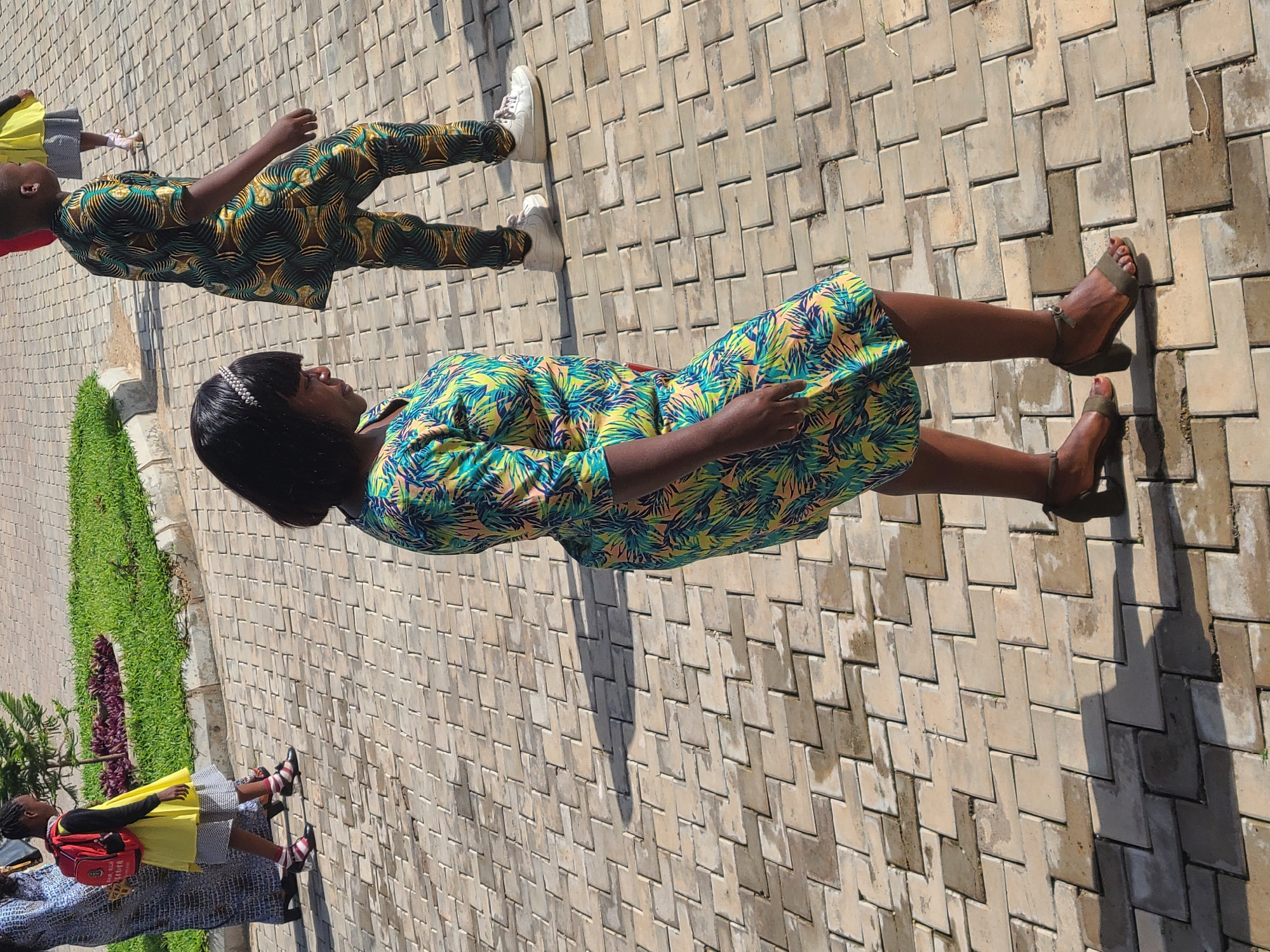 A brightly lit outdoor scene shows four people standing on a paved surface next to a patch of grass. One person is wearing a dress patterned with blue, yellow, and green colors, and sandals. Another person is wearing a matching top and pants with black, yellow, and green patterns, and white sneakers. Two other people are present but partially out of frame, one wearing a yellow top, black jacket and skirt, and the other wearing a light blue top, and black and white skirt. The setting appears to be a pedestrian area within a city.