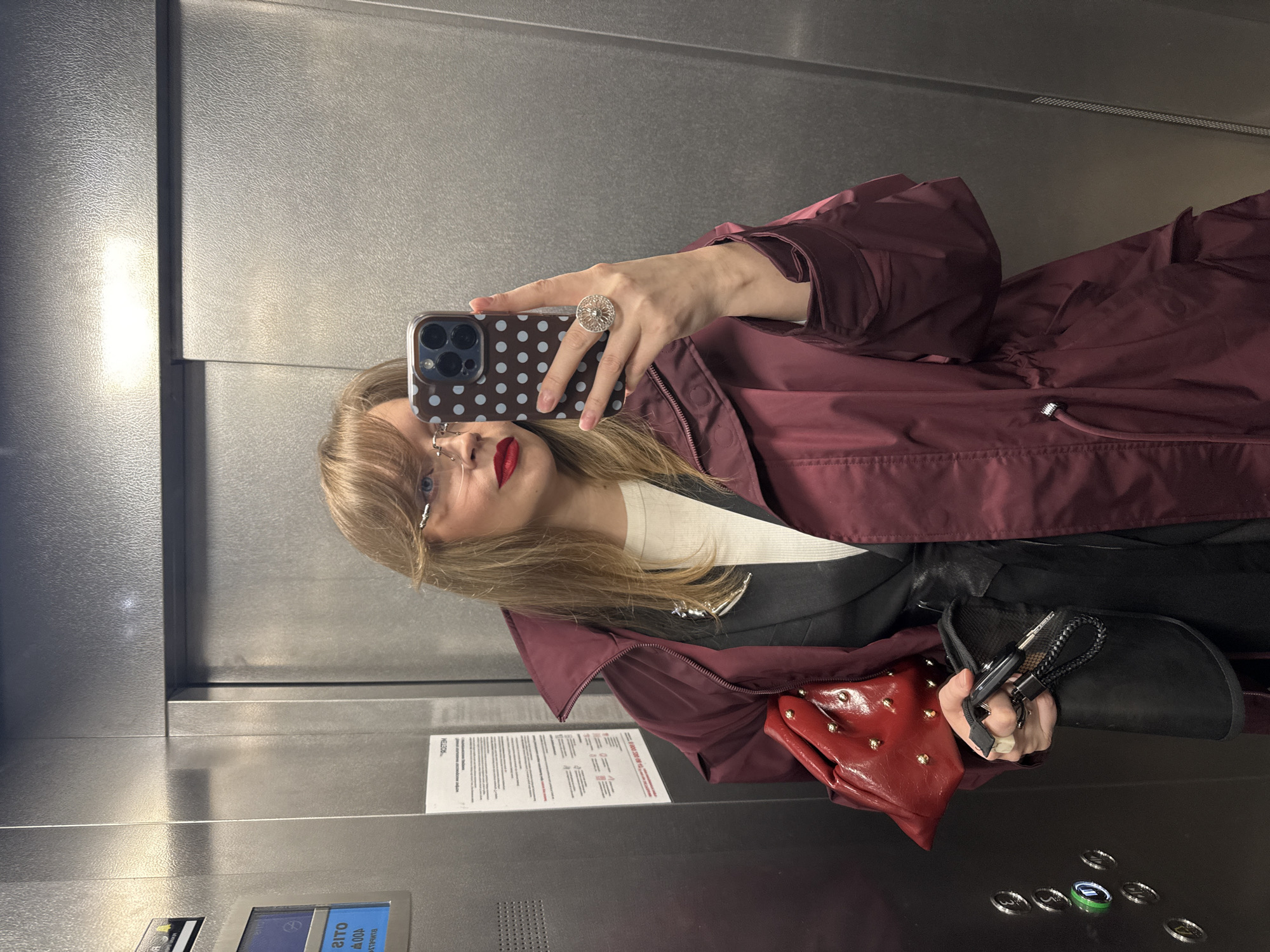A woman is taking a selfie in an elevator. She is wearing a burgundy jacket and red lipstick. She is holding a red purse and a black shoe. Her phone has a polka dot case. There is a notice posted on the elevator wall with text in Russian. Based on the text visible on the elevator control panel, the location is Izhevsk, Russia.