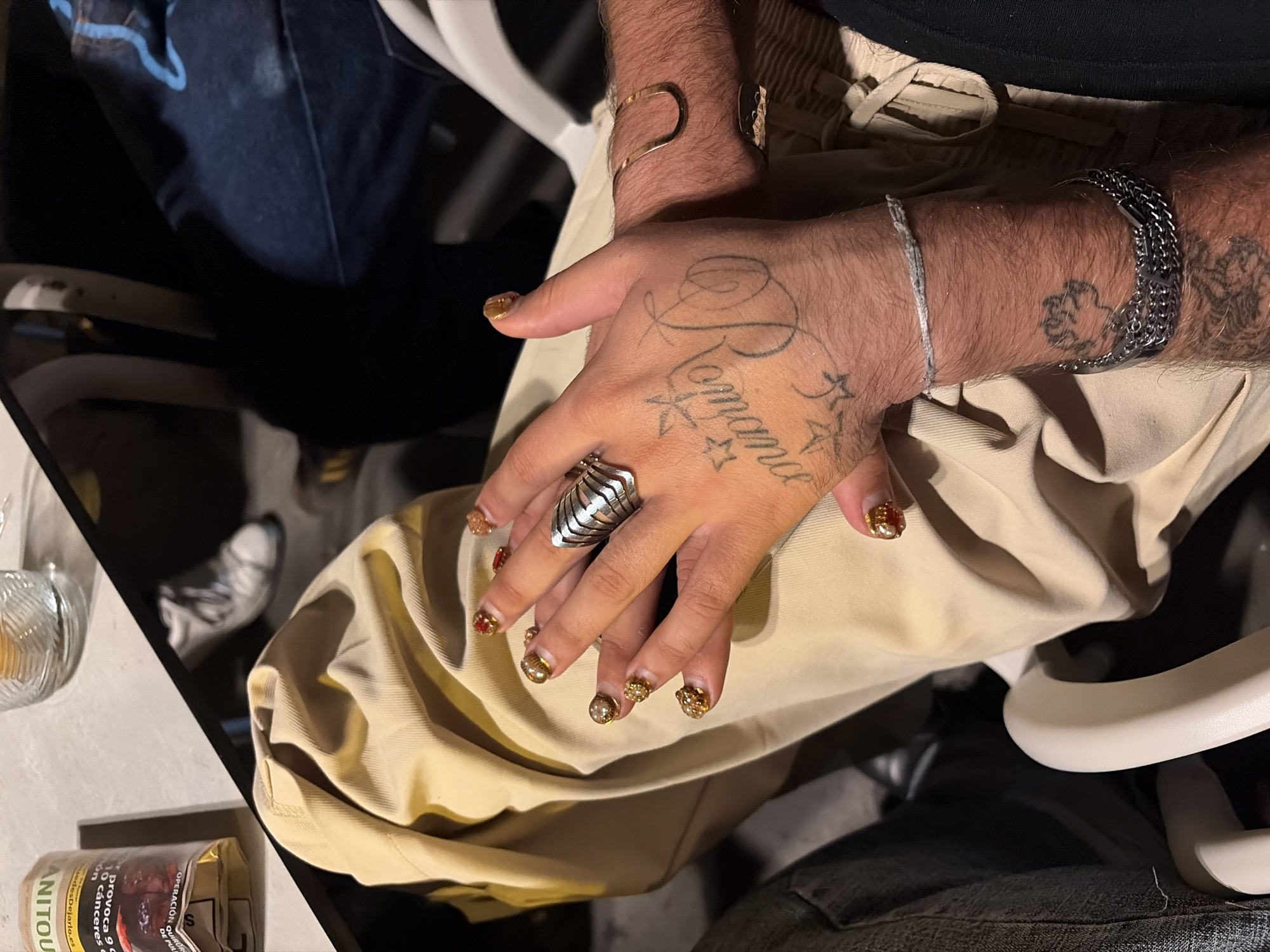 The image shows a person's hands resting on their lap. The hands are adorned with jewelry: a large silver ring on the left hand, and bracelets on both wrists. One hand is tattooed with the word "Romance" and several stars. The person is wearing a gold-colored bracelet. The person is also wearing manicured nails with gold and red embellishments. The person is wearing light-colored pants and sits on a white chair. A pack of "Canitou" cigarettes can be seen on a white table next to the chair. The environment appears to be indoors. There are no visible clues suggesting that the image was taken specifically in Barcelona, Spain.