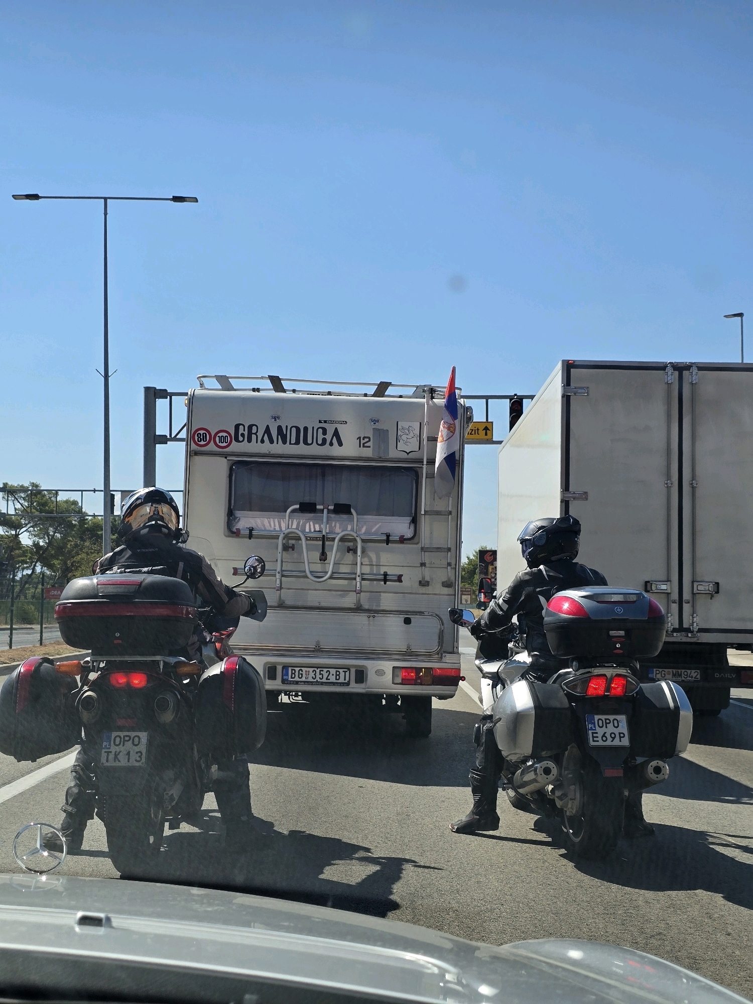 The image shows a sunny day with a clear blue sky, likely on a highway, with two motorcyclists in the foreground. The motorcyclist on the left is riding a dark-colored motorcycle with red saddlebags and a red top case. The motorcyclist is wearing a black leather jacket and pants, and a helmet with a reflective visor. The motorcycle's license plate is visible and reads "PL OPO TK13".
In front of this motorcycle is a white camper van with the text "GRANDUCA 12" on the back. A Serbian flag is mounted on the side of the camper. The camper has various stickers, including speed limit indicators of 80 and 100. A license plate on the camper reads "BG 352-BT".
To the right of the camper is a large, white truck. The second motorcyclist is on a silver and black motorcycle, also wearing a black leather outfit and a helmet. This motorcycle has silver saddlebags and a black top case. The license plate of this motorcycle reads "PG E69P".
The overall scene suggests a road trip or travel. The presence of road signs and the highway setting indicate a transportation route. The bright sunlight casts shadows on the road. The image does not provide specific visual cues to pinpoint a location within the city of Podgorica, Montenegro, but the license plates suggest travel from or within Serbia and Montenegro.