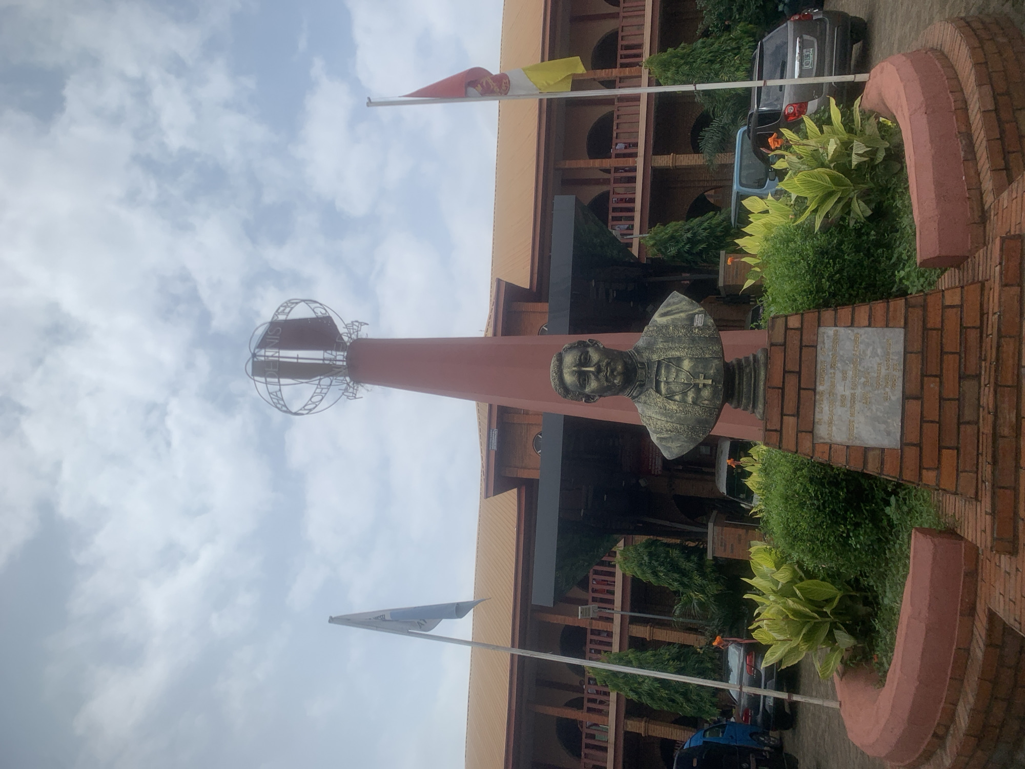 The image depicts the exterior of the Archbishop Dennis Memorial High School in Onitsha, Nigeria. The main subject is a bronze bust of a man in clerical attire, placed on a brick pedestal. Behind the bust, a tall red tower stands, topped with a metallic globe-like structure. Two flags are visible: one on the left, a light blue and white flag, and another on the right, a red and white flag with a crest. A large, two-story brick building with a brown roof forms the backdrop. The building features arched windows and a covered walkway with railings. Greenery, including trees and manicured bushes, surrounds the bust. Several cars are parked in front of the building. The sky is overcast with scattered clouds. A plaque is visible on the brick pedestal in front of the bust, containing engraved text.