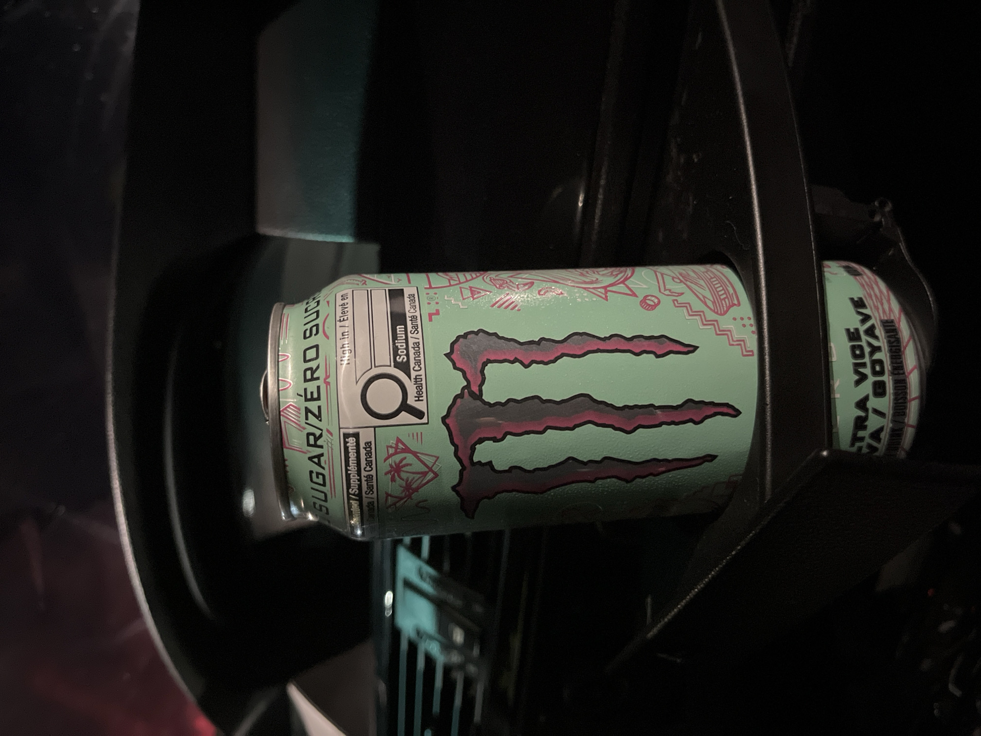 A can of Monster Energy drink, specifically the "Ultra Violet" flavor, is sitting in a car's cupholder. The can is primarily light green with pink and black designs. The label includes nutritional information in English and French, with "SUGAR/ZÉRO SUCRE" prominently displayed. The cupholder is black, and the car's interior is generally dark.
There is no indication of the location being Shumen, Bulgaria.