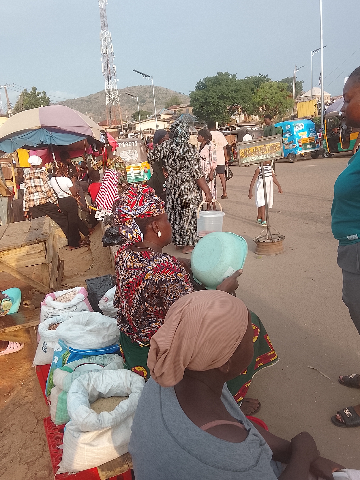 **Location:** Gwong, Nigeria, likely a busy market area.
**People:** Several women, some seated selling goods, some standing and walking. A young girl is also present.
**Subjects:** Market vendors, customers, pedestrians.
**Objects:** Sacks of goods (likely grains or beans) for sale, buckets, a sign propped up by a tire rim, a three-wheeled vehicle (tricycle) with advertising.
**Actions:** Selling, buying, walking, observing.
**Interactions:** Vendors interacting with customers, people navigating the busy market area.
**Environment:** Outdoor market setting with stalls, tents, and pedestrian traffic on a paved road. A telecommunications tower is visible in the background, suggesting a populated area.