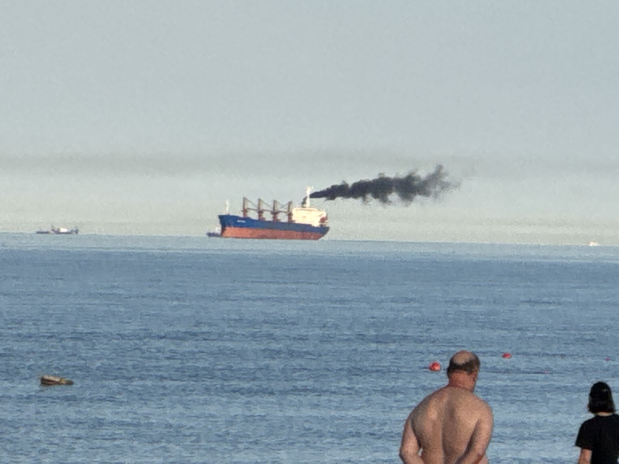 The image depicts a coastal scene in Novorossiysk, Russia, looking out over a body of water, likely the Black Sea. In the foreground, with their backs to the camera, are two individuals observing the sea. One is a shirtless man with a visible bald spot, and next to him is another person with dark hair wearing a dark t-shirt.
The central focus of the image is a large cargo ship, possibly a bulk carrier, in the mid-distance. The ship has a red hull, a blue upper structure, and a white cabin and funnel area. It is actively moving and emitting a prominent plume of thick, black smoke from its funnel, which drifts towards the right of the frame. Faint text, possibly the ship's name "COMA," can be seen on its bow.
Further in the distance, on the horizon line, several smaller boats or vessels are faintly visible. Closer to the foreground, a piece of debris or a log floats on the water's surface to the left. Additionally, several red buoys are scattered across the mid-ground water.
The time of day appears to be daytime, with clear skies and good visibility. The water is relatively calm. The scene captures a typical day at a waterfront, with people observing maritime activity. The black smoke from the ship is a notable detail, highlighting its emissions.