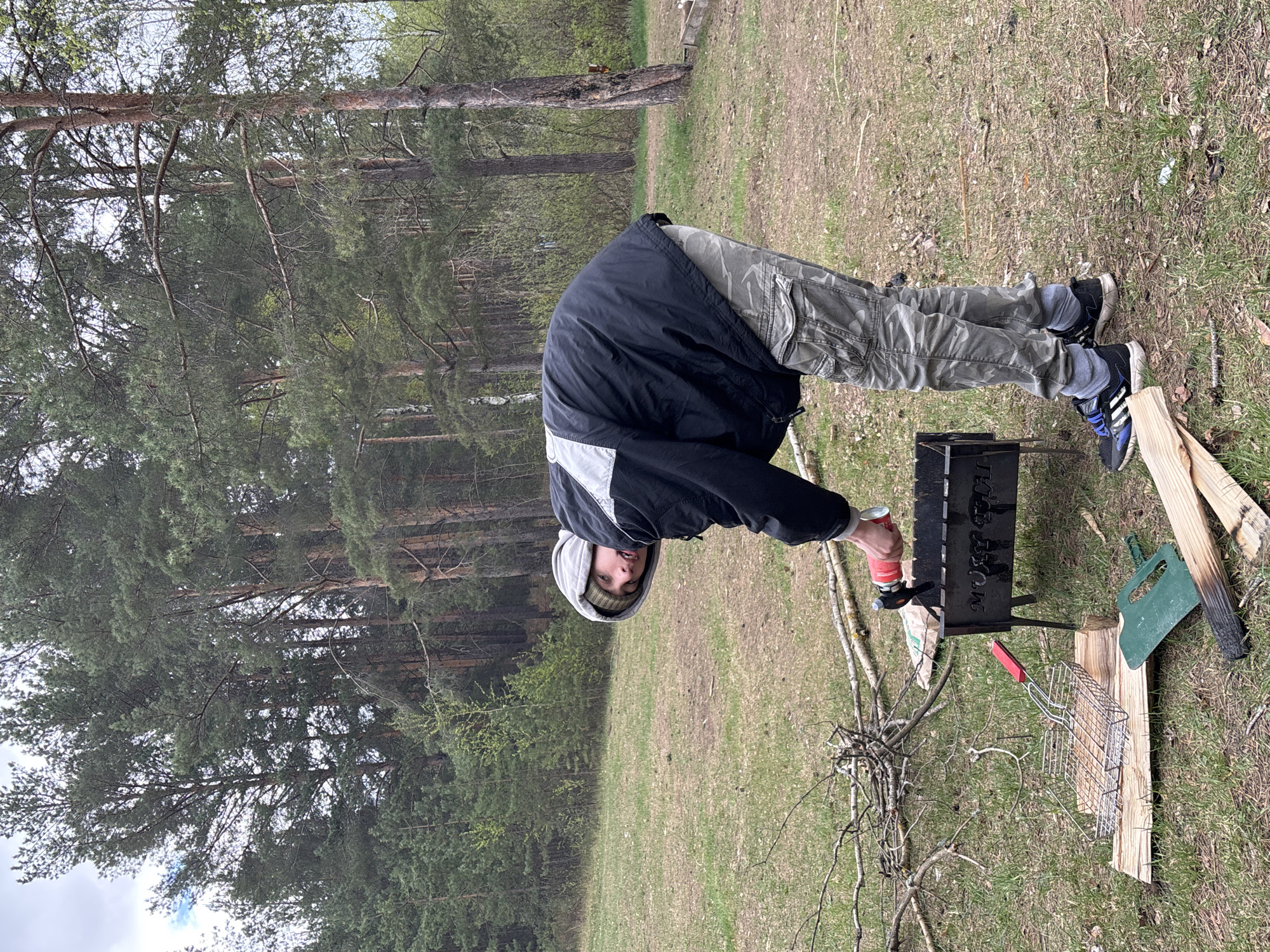 A person is setting up a barbecue in a grassy area near a forest in Vekoshinka, Russia. The person is wearing a black jacket with a gray hood, camouflage pants, and blue and black sneakers. They are bending over a black metal barbecue grill, holding a red and white canister. To the right of the grill, there is a piece of wood with a grilling basket and a green plastic container. There are small branches on the ground near the grill. Behind the person is a dense forest with tall trees. The sky is overcast.