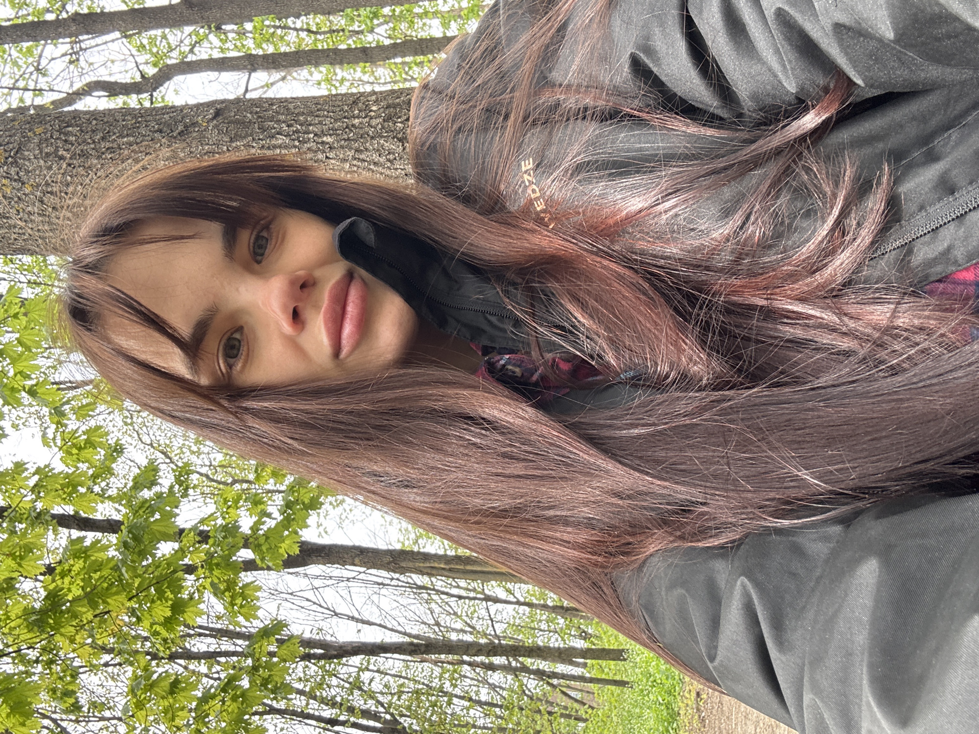 A young woman with long brown hair, wearing a dark jacket, is in a wooded area in Rostov-on-Don, Russia. The background shows trees with green leaves and other trees with bare branches. The woman is facing the camera and appears to be taking a selfie.