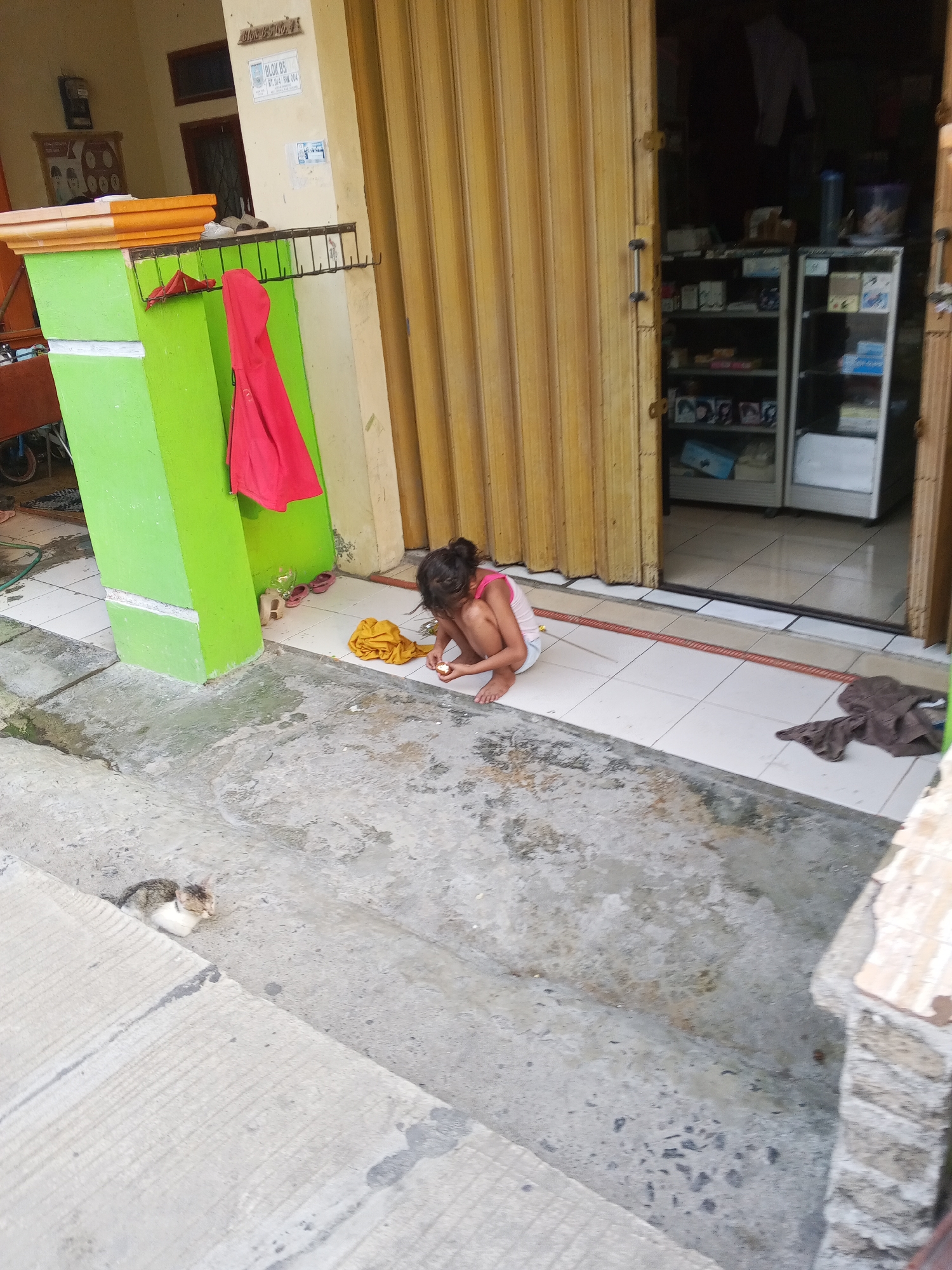 A young girl in a pink tank top and white shorts is crouching on a tiled floor outside a shop in Ciruas, Indonesia. She is holding a small, light-colored object in her hands and appears to be examining it. In front of her is a pile of yellow fabric. To her left, a red garment hangs from a rack attached to a bright green pillar. A brown cloth lies on the floor to her right. Further to the left, a small cat with grey and white fur is resting on a concrete step. Behind the girl, a set of yellow, corrugated metal doors are partially open, revealing shelves stocked with various items inside the shop. The environment appears to be a street or alleyway in a residential or commercial area.