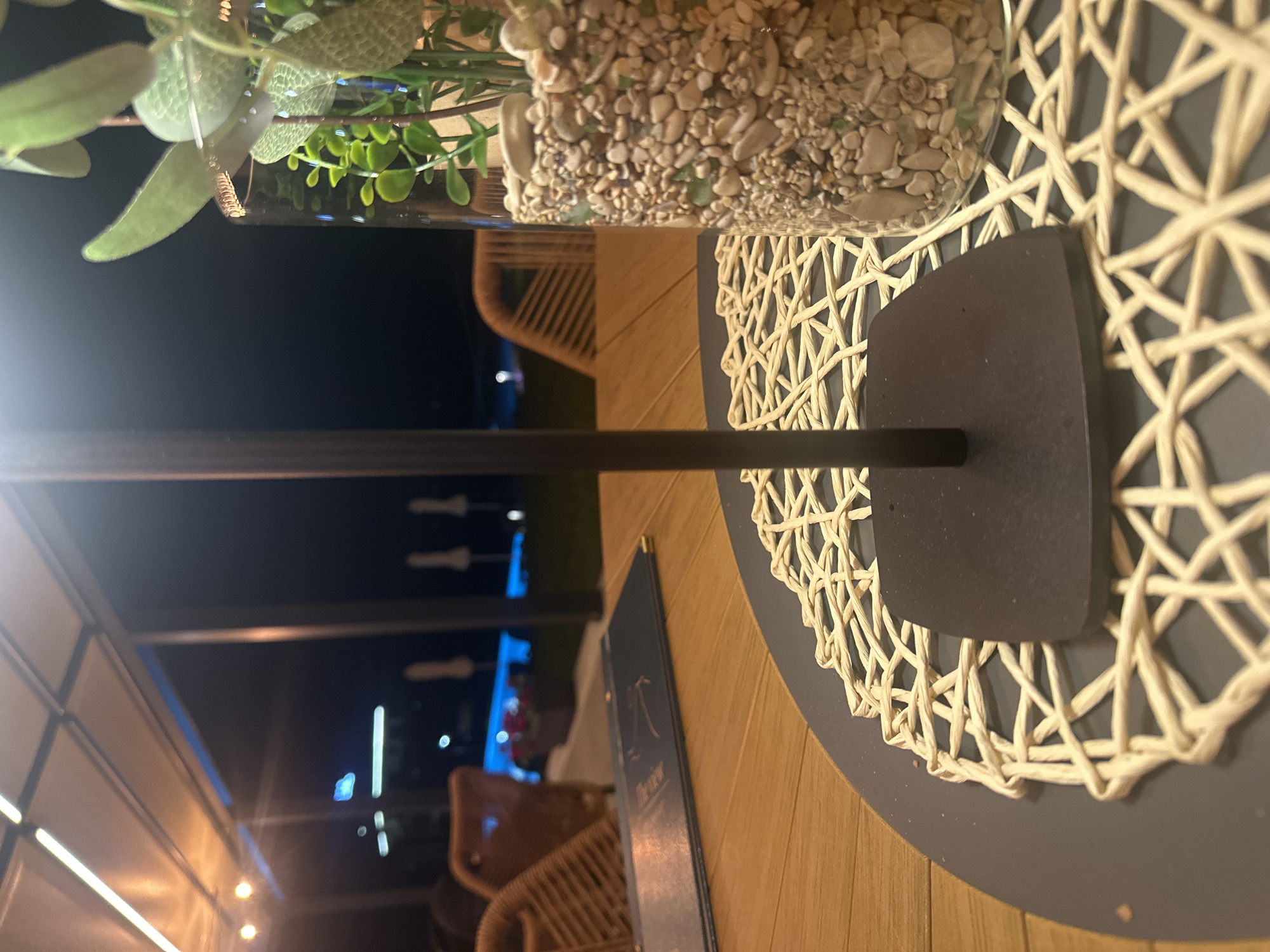 The image shows an outdoor seating area at night. A glass vase filled with small stones and artificial plants sits on a table. A dark metal stand supports the vase. A woven placemat is underneath the stand. The table is made of wood. In the background, there are wicker chairs and some lights. The scene appears to be a restaurant or cafe. The time of day is night, and the weather is not discernible. No people are visible in the image. There is some text on a dark blue menu lying on the table, but it is largely illegible due to the angle and lighting.