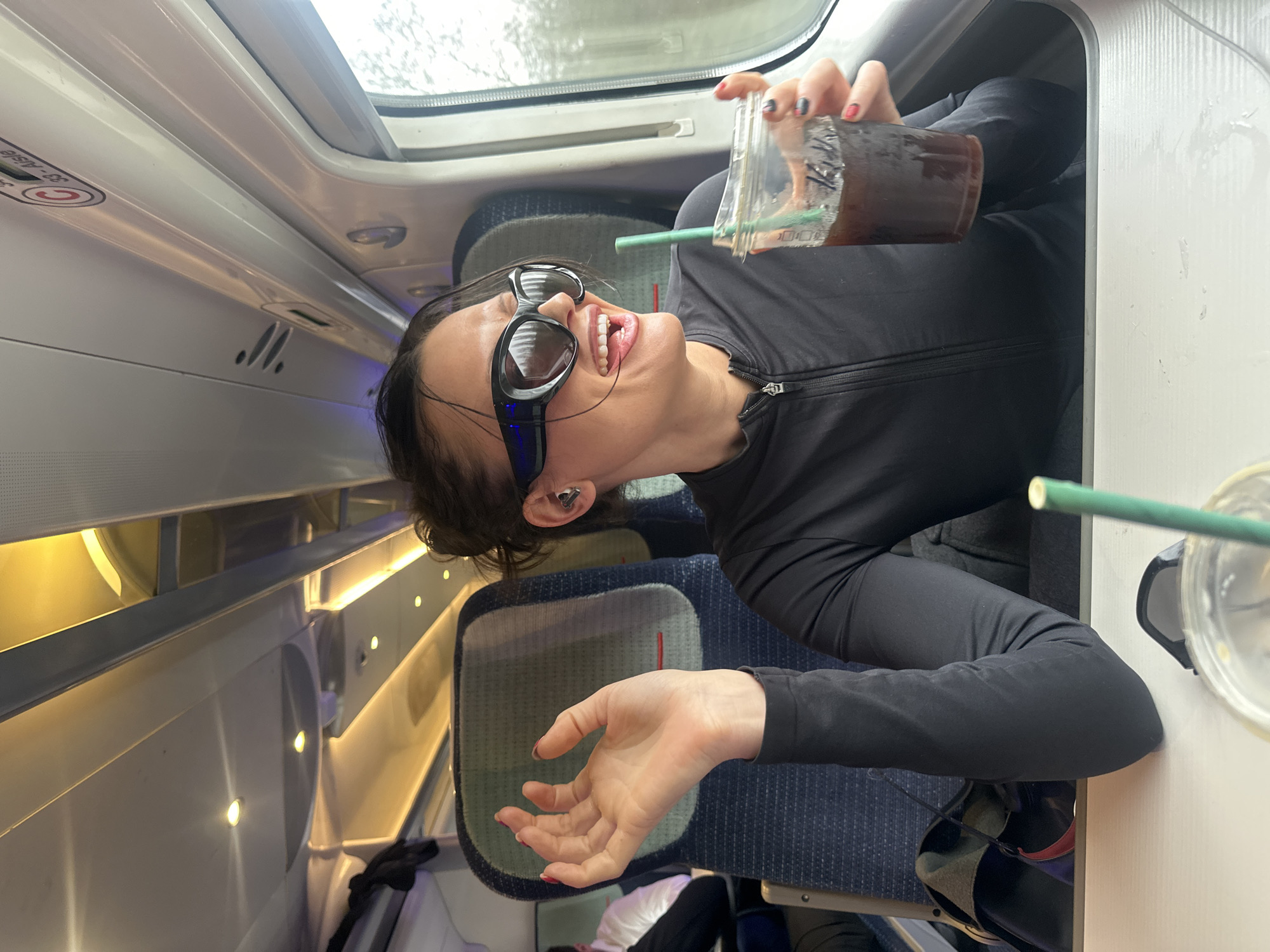 The image depicts the interior of a modern LNER Azuma train carriage, likely operating on the East Coast Main Line to or from London King's Cross station, London, United Kingdom. A female subject is seated by a window, facing right and partially away from the camera. She wears a black long-sleeved, quarter-zip top, large black sports-style sunglasses, and a light-colored earbud in her right ear. Her dark hair is styled back. Her mouth is open in an animated expression, with her tongue slightly extended. She holds a clear plastic cup containing a dark, iced beverage with a green straw in her right hand. Her left hand is raised, palm open, with fingers slightly splayed. Both hands display red and black-patterned nail polish. In front of her is a white table surface with visible liquid condensation marks. The train's blue fabric seats with green dots and a red stripe are visible. Overhead, recessed spotlights and linear ambient lighting illuminate the compartment, and an overhead sign indicates "C 33 - Aisle". Through the window, an indistinct, blurred grey-green exterior is visible, characteristic of a moving train. A male passenger in a white shirt and dark clothing is partially visible in the seat behind and to the left of the female subject.