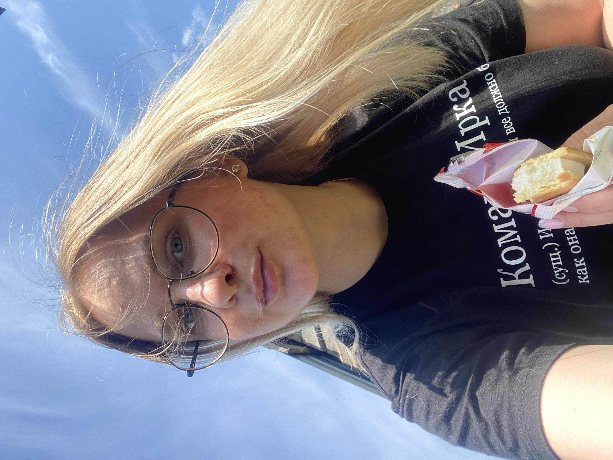 In Kauchuk Stantsiya, Russia, a young woman with blonde hair and glasses is taking a selfie outdoors. She is wearing a black t-shirt with white text. She is holding a small pastry wrapped in red and white packaging, with one bite taken out of it. The background is a clear blue sky with a few wispy clouds. The lighting suggests it is a sunny day.