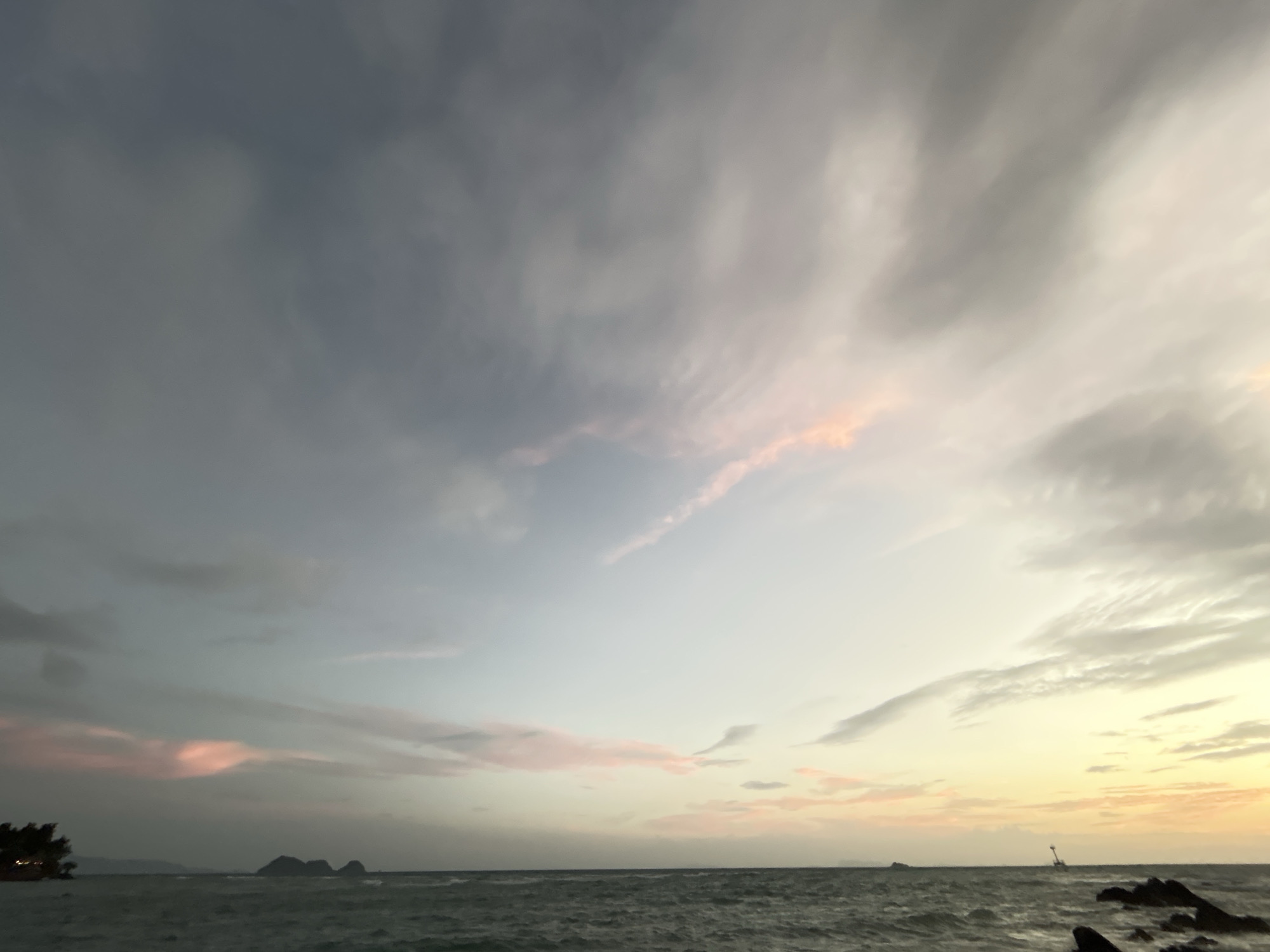 The image captures a serene ocean view at dusk, likely in Ko Pha-ngan District, Thailand. The sky is a canvas of soft hues, transitioning from a dusky blue in the upper left to lighter shades of grey, pink, and a warm yellow glow towards the right horizon. Wispy clouds stretch across the sky, some tinged with pink from the setting sun.
The ocean in the foreground is a deep, choppy grey, with visible waves suggesting a gentle breeze. The waterline is marked by dark, jagged rocks on the right side of the frame, while the left side shows a distant, dark silhouette of land with some vegetation and structures. Further out in the ocean, a few small islands or rock formations are visible, along with a solitary, slender structure that appears to be a navigational marker or a very small boat in the distance. There are no people or animals visible in the frame. The overall impression is one of a tranquil, natural landscape at the end of the day.