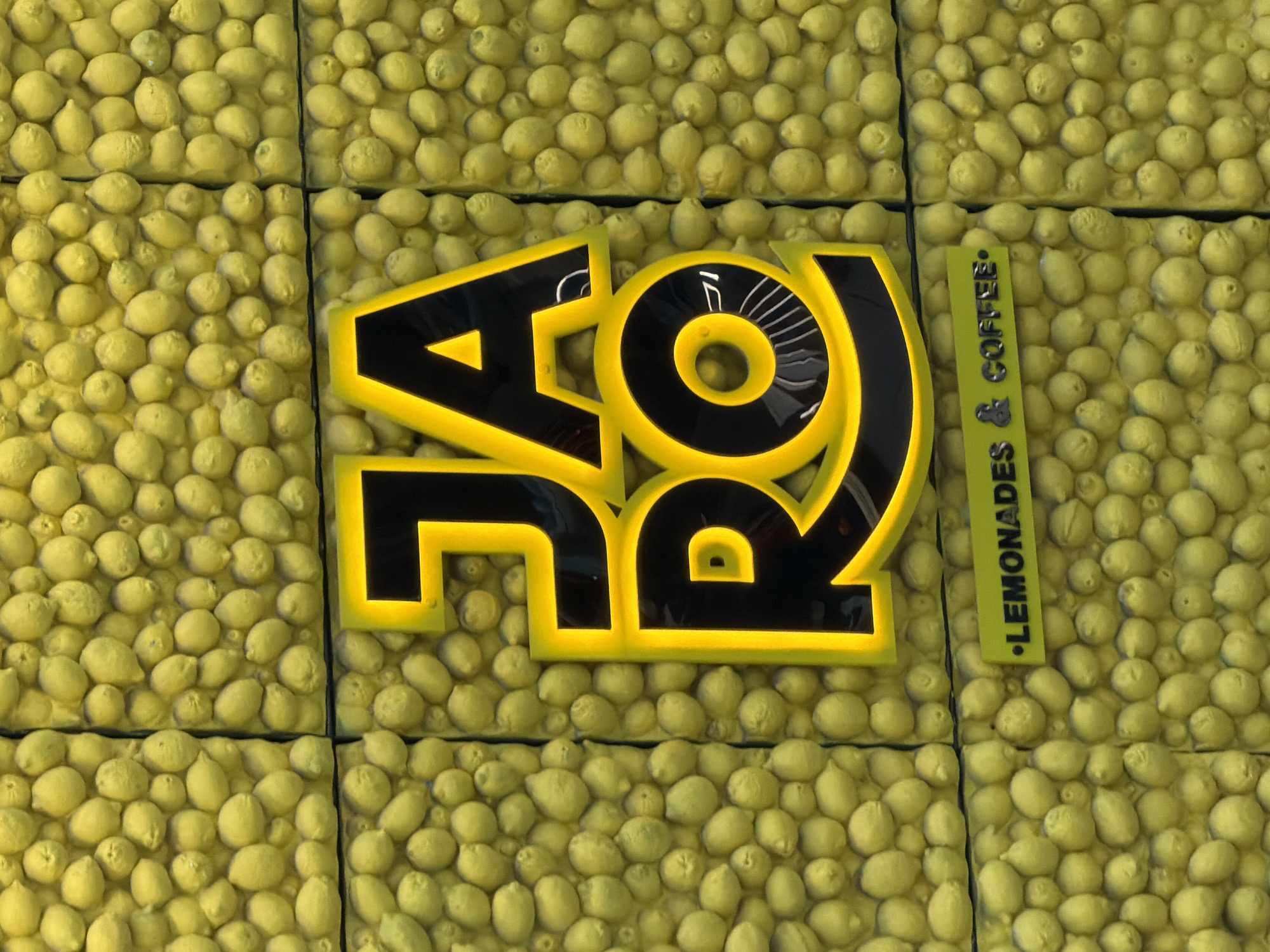 This is a close-up, eye-level, vertical, medium shot of a sign for "JARO LEMONADES & COFFEE" that is mounted on a wall covered in artificial lemons. The sign is illuminated with yellow neon lighting around black lettering. The letters "JA" are stacked on top of "RO". Below the main sign, a horizontal green banner with white text reads ". LEMONADES & COFFEE .". The wall behind the sign is densely packed with small, yellow, artificial lemons that create a textured surface. The lighting in the image is bright, highlighting the neon sign and the lemons. There are no people visible in the image. The setting appears to be an outdoor or indoor commercial space.
