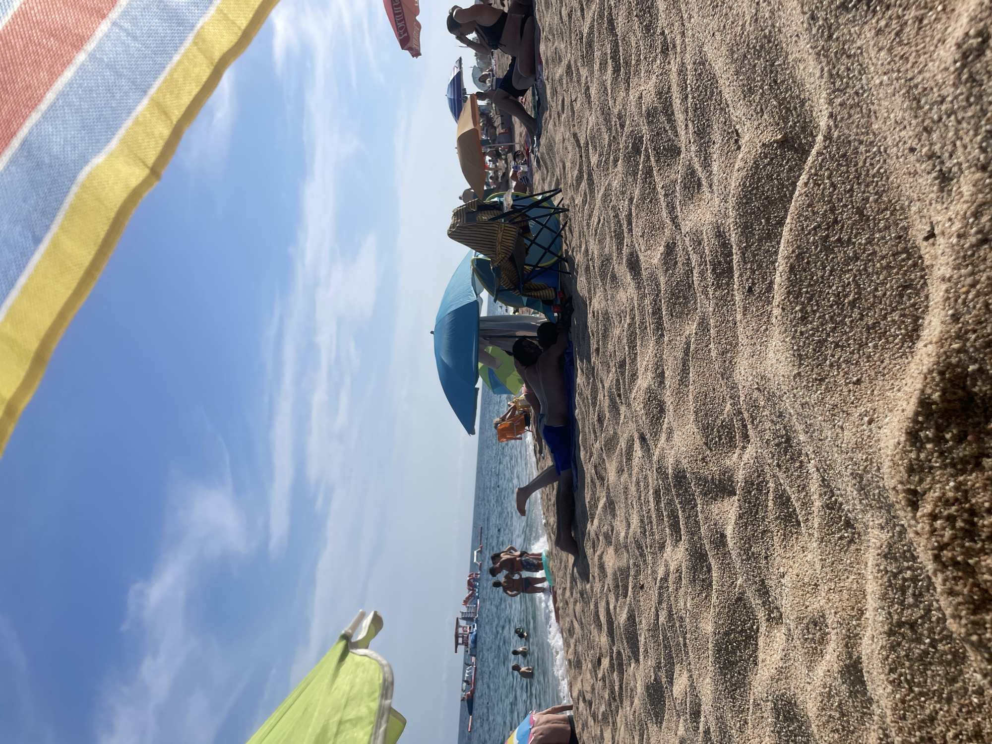 The image depicts a sunny day at the beach in Canet de Mar, Spain. In the foreground, the rippled sand dominates the frame, with sunlight casting shadows and highlighting the texture. A person wearing blue swim trunks is lying on their stomach in the sand, facing away from the camera, with their legs extended towards the water.
In the middle ground, the sea is visible with gentle waves lapping at the shore. Several people are in the water, some standing and others swimming. Further out in the sea, there are inflatable water attractions. Various beach umbrellas are scattered across the sand, providing shade. A blue umbrella is prominent, with a person sitting under it. To the right, another beachgoer is seated on the sand, wearing a dark bikini and adjusting their hair. Other beach umbrellas and people are visible in the background, contributing to a lively beach atmosphere. The sky above is a clear blue with scattered white clouds. The scene suggests a typical summer day at a popular beach.