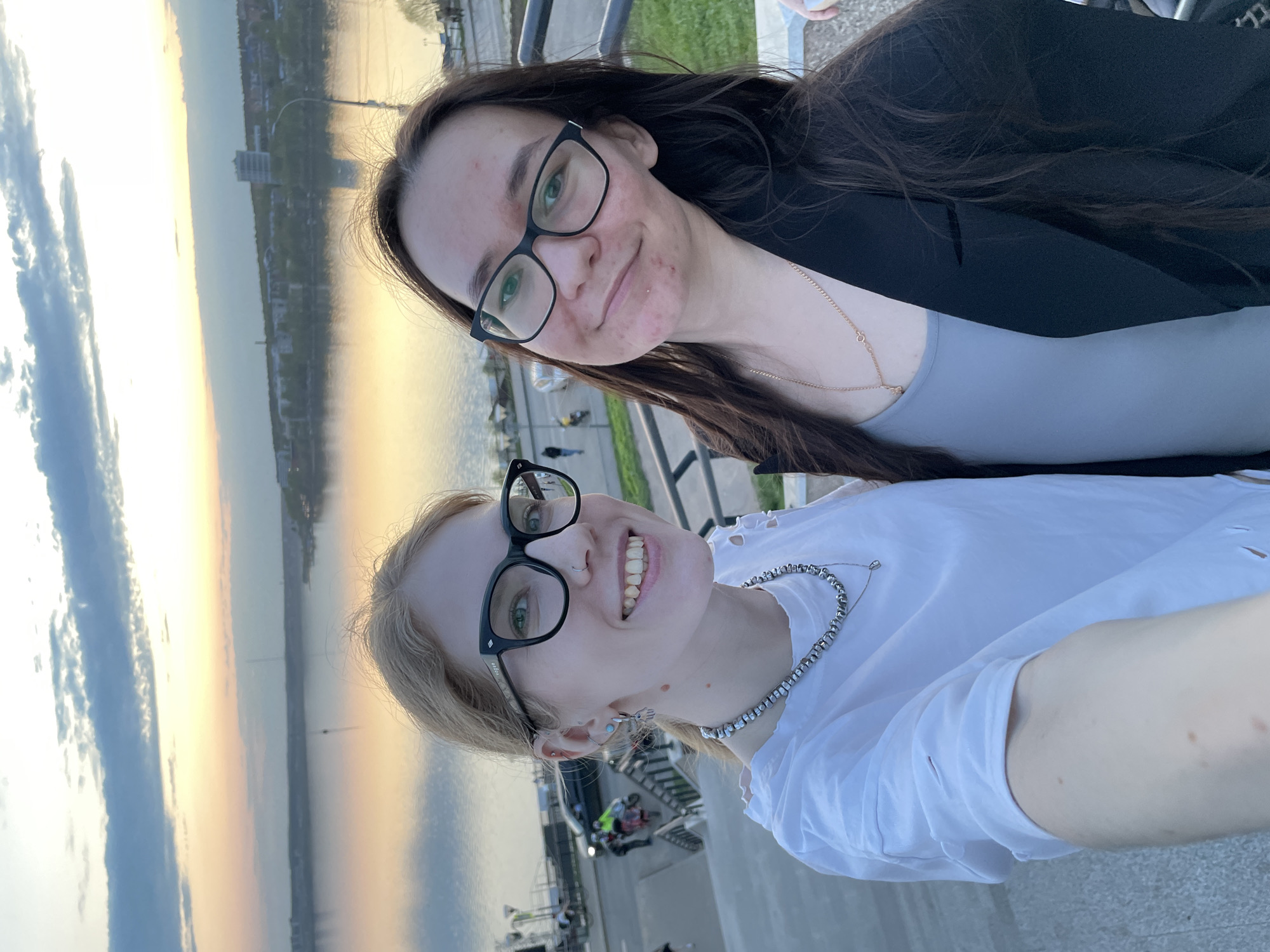 Two young women pose for a selfie outdoors during sunset. The woman in the foreground has blonde hair and wears black-framed glasses, a white t-shirt, and a silver beaded necklace. The woman in the background has long brown hair and wears black-framed glasses, a gray top, and a black blazer. A delicate gold necklace is visible on her chest. The background shows a river reflecting the colors of the sunset, with urban structures along the shoreline. The picture was taken in Izhevsk, Russia.